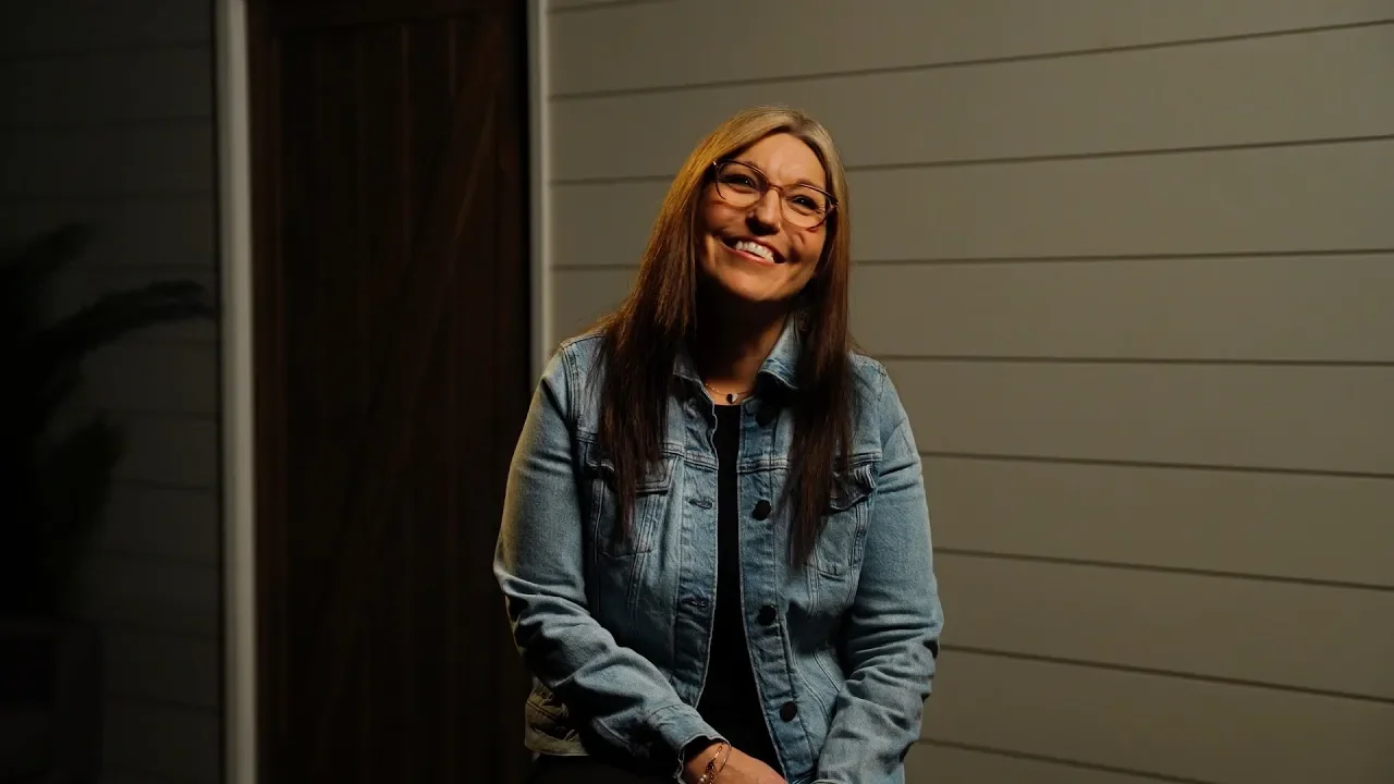 A woman with long brown hair, glasses, and a denim jacket smiling at the camera, standing in front of a beige horizontal siding wall at night.