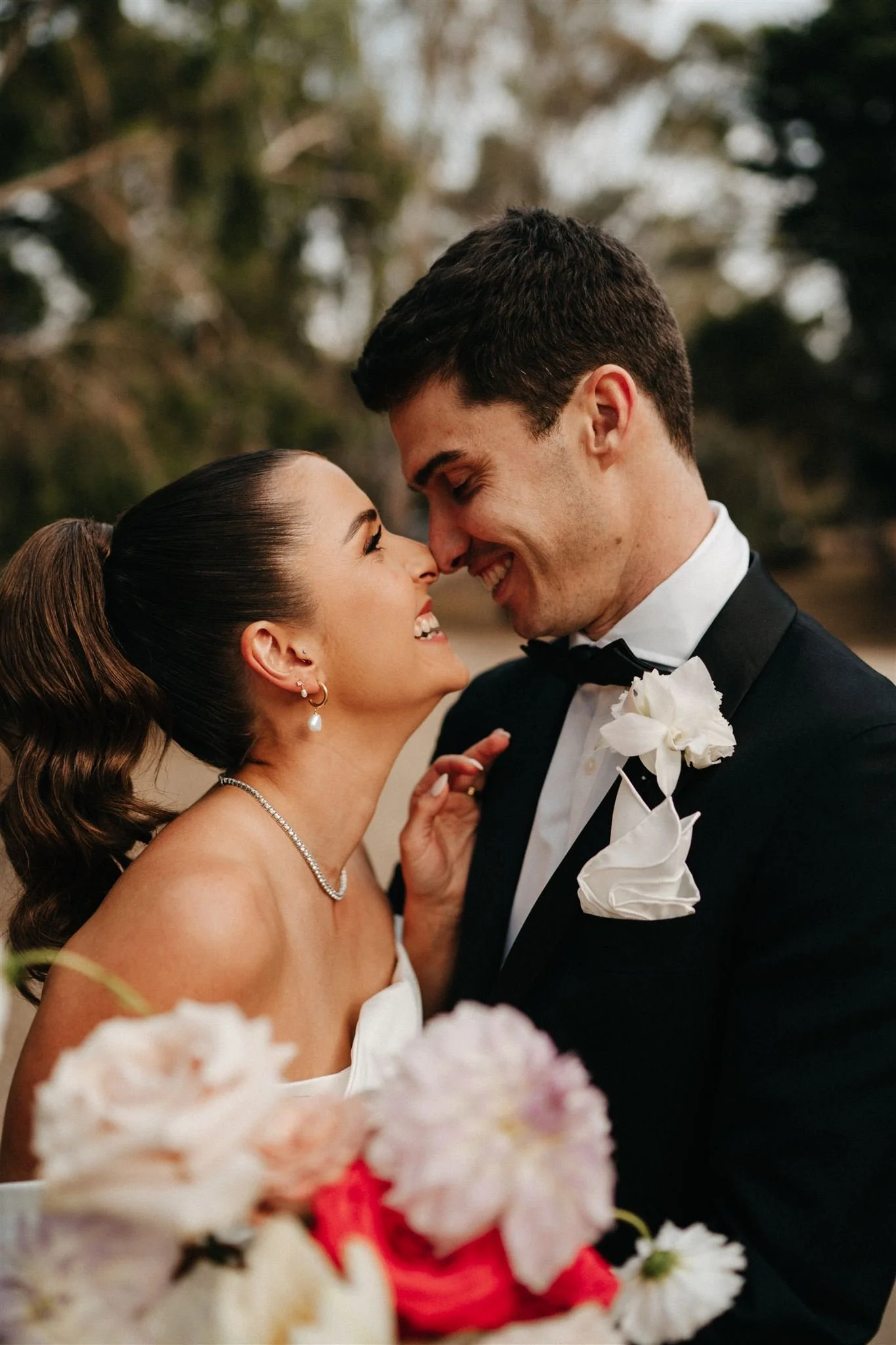 A bride and groom smiling closely, their foreheads touching, during a wedding celebration outdoors with blooming flowers in the foreground.