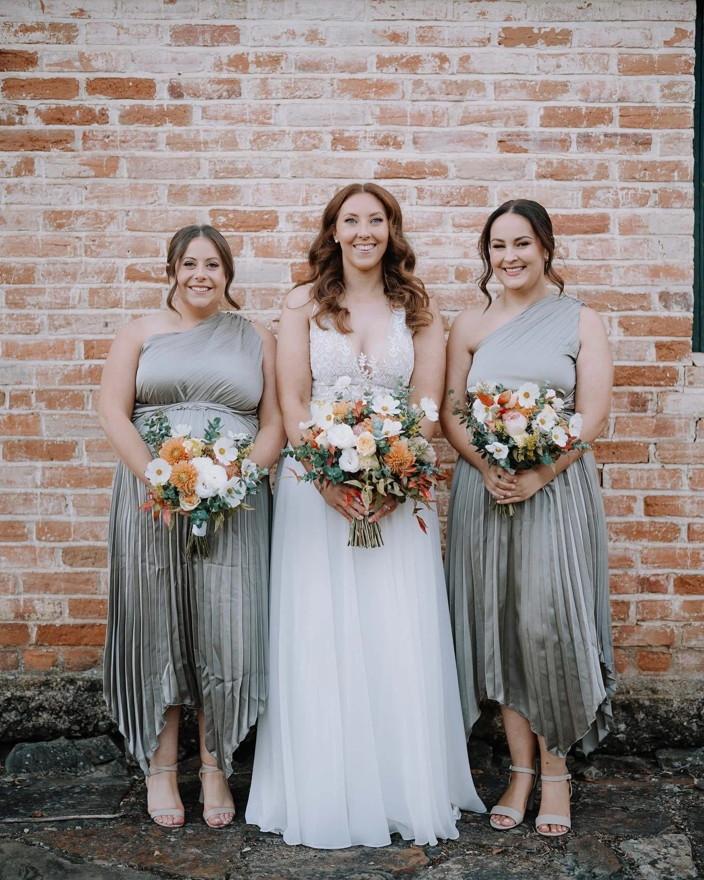 Bride in white dress with two bridesmaids in gray dresses holding bouquets of flowers, standing in front of a brick wall.