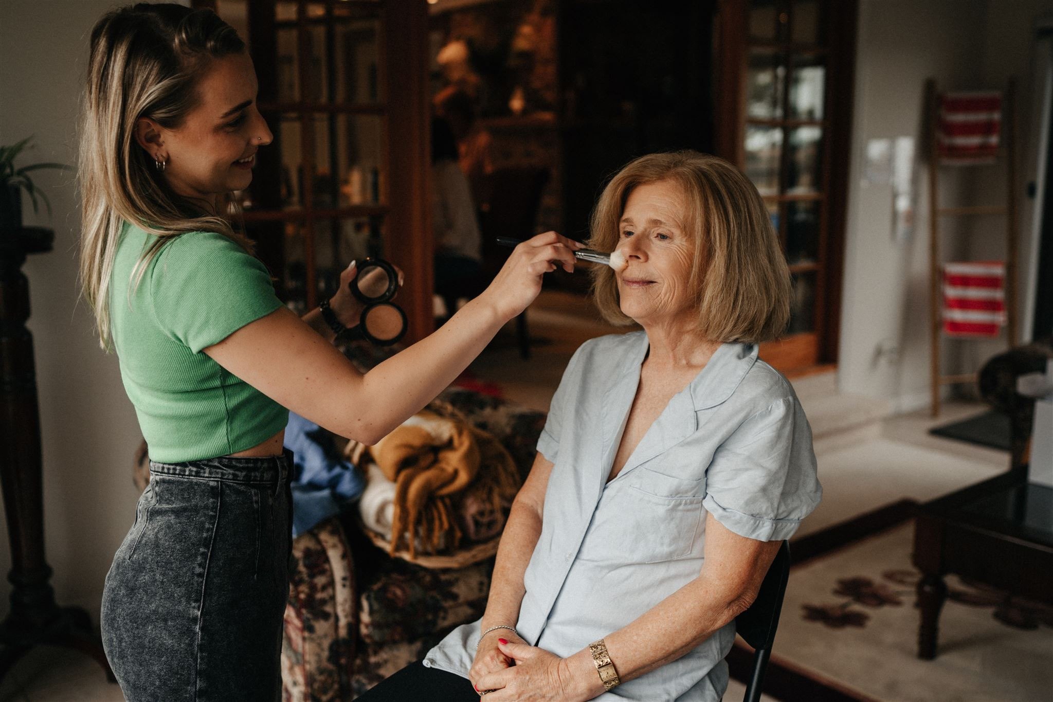 A young woman applying makeup to an older woman using a brush, with the older woman sitting and smiling in a cozy, warmly-lit living room.