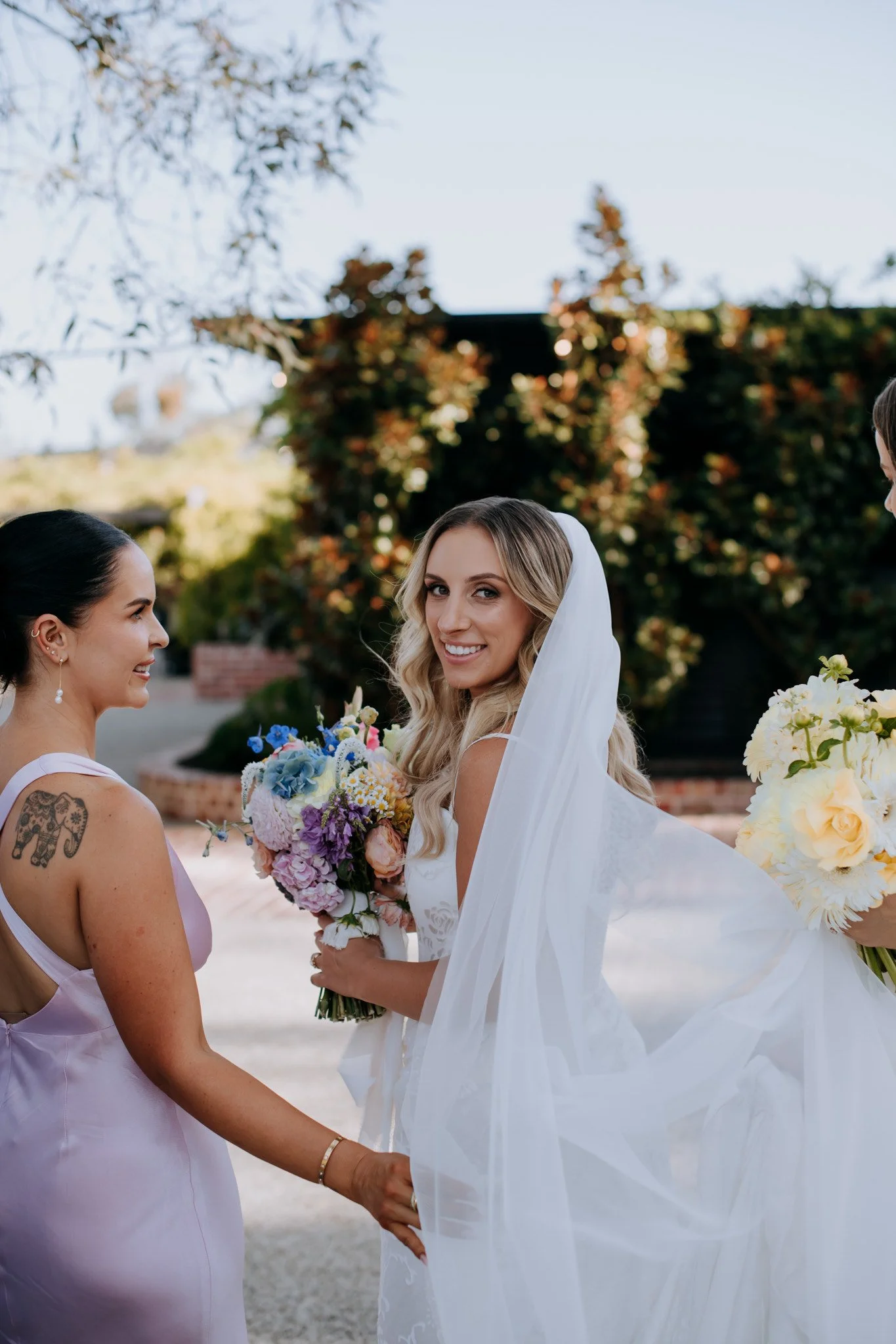 A bride in a white wedding dress and veil holding a bouquet of flowers, smiling at her wedding party outside with trees and blue sky in the background.