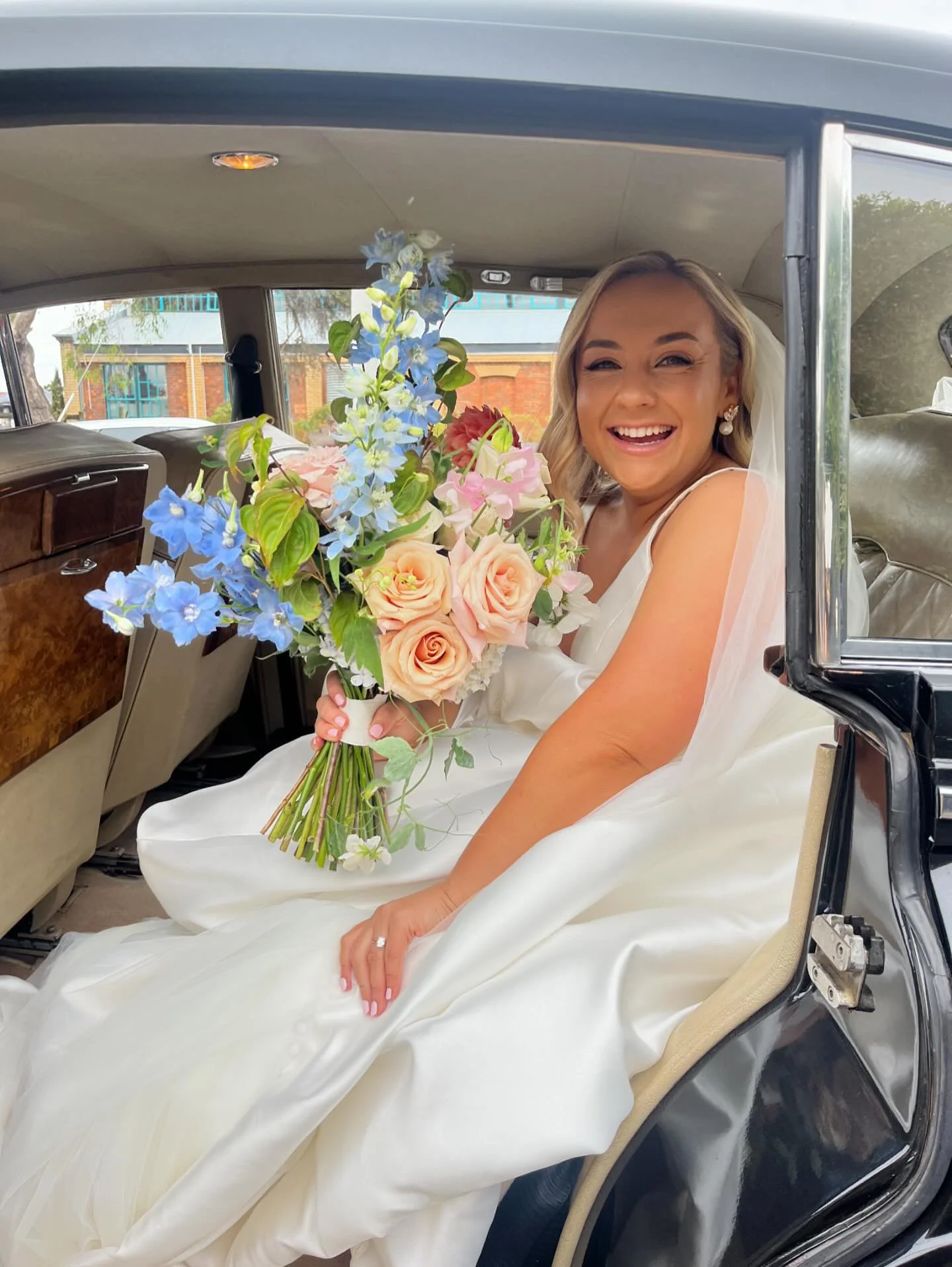 A bride sitting inside a vintage car, smiling and holding a large bouquet of colorful flowers, wearing a wedding gown and veil.