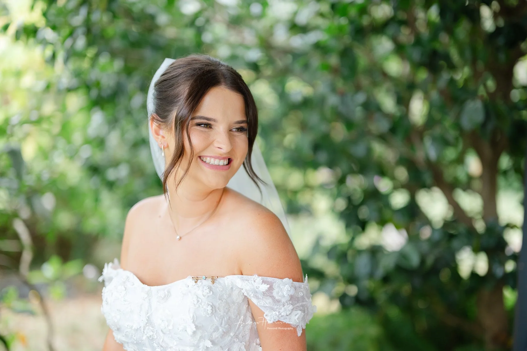 A smiling bride with dark hair styled in loose waves, wearing a white off-the-shoulder wedding dress with lace details, standing outdoors with a background of green trees.