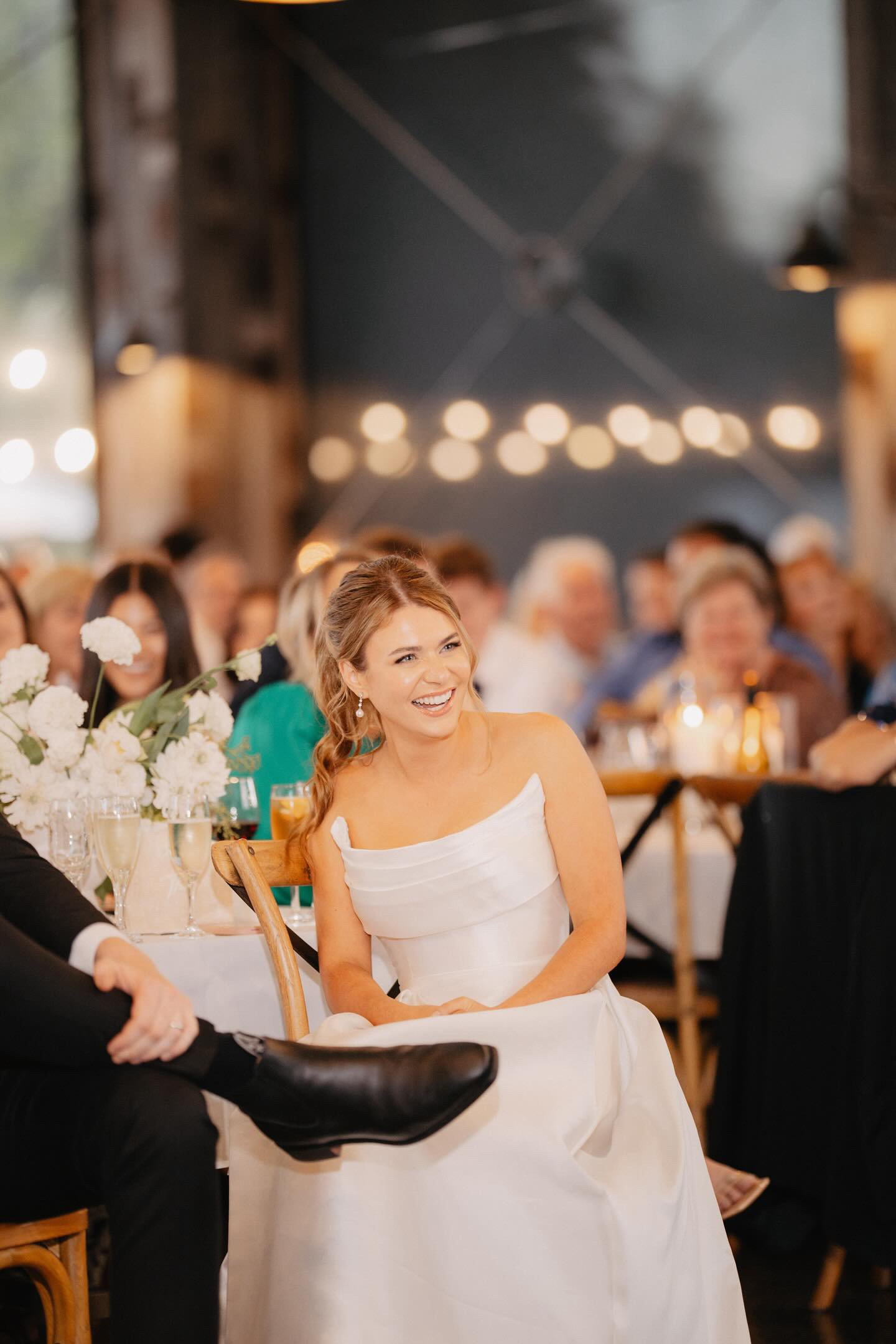 A smiling woman in a white off-the-shoulder dress sitting at a wedding reception surrounded by guests, with flowers and table settings visible in the background.