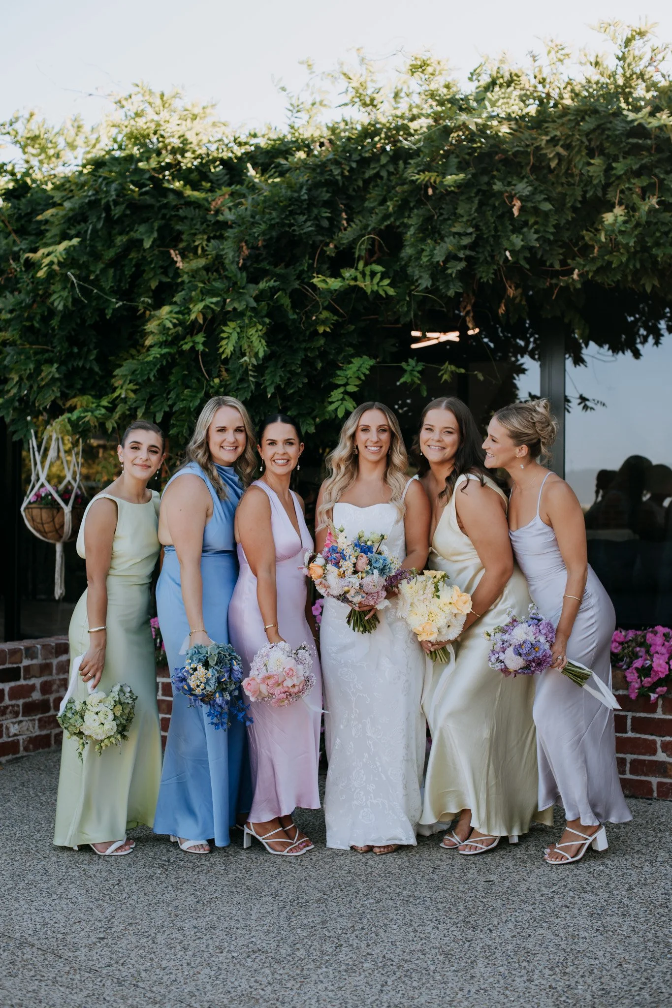 Group of six women in pastel dresses holding bouquets, standing outdoors in front of greenery and flowers, smiling at the camera.