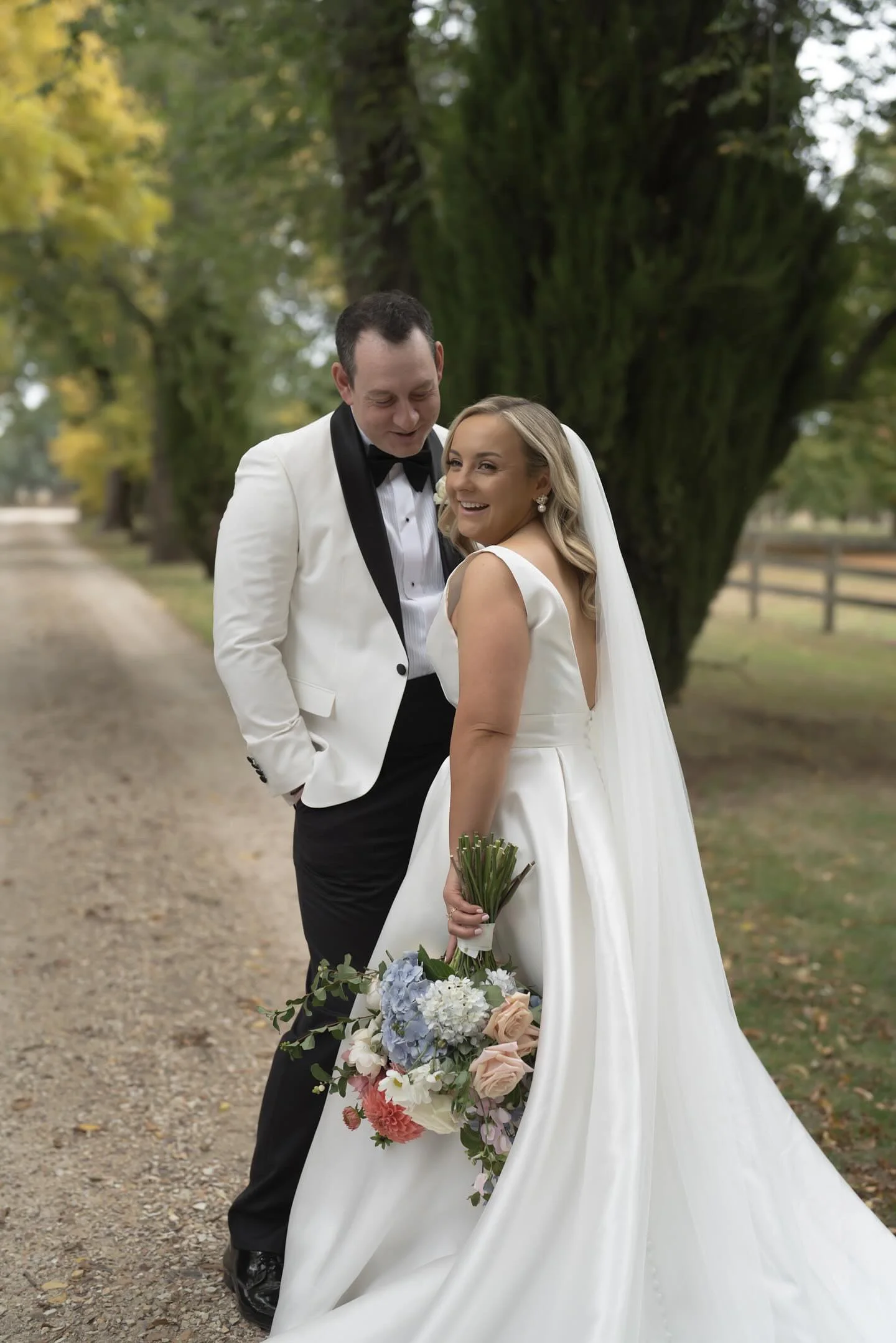 A bride and groom smiling and standing close together outdoors, surrounded by trees, during their wedding photo. The bride is holding a bouquet of flowers, and the groom is wearing a tuxedo with a white jacket and black bow tie.