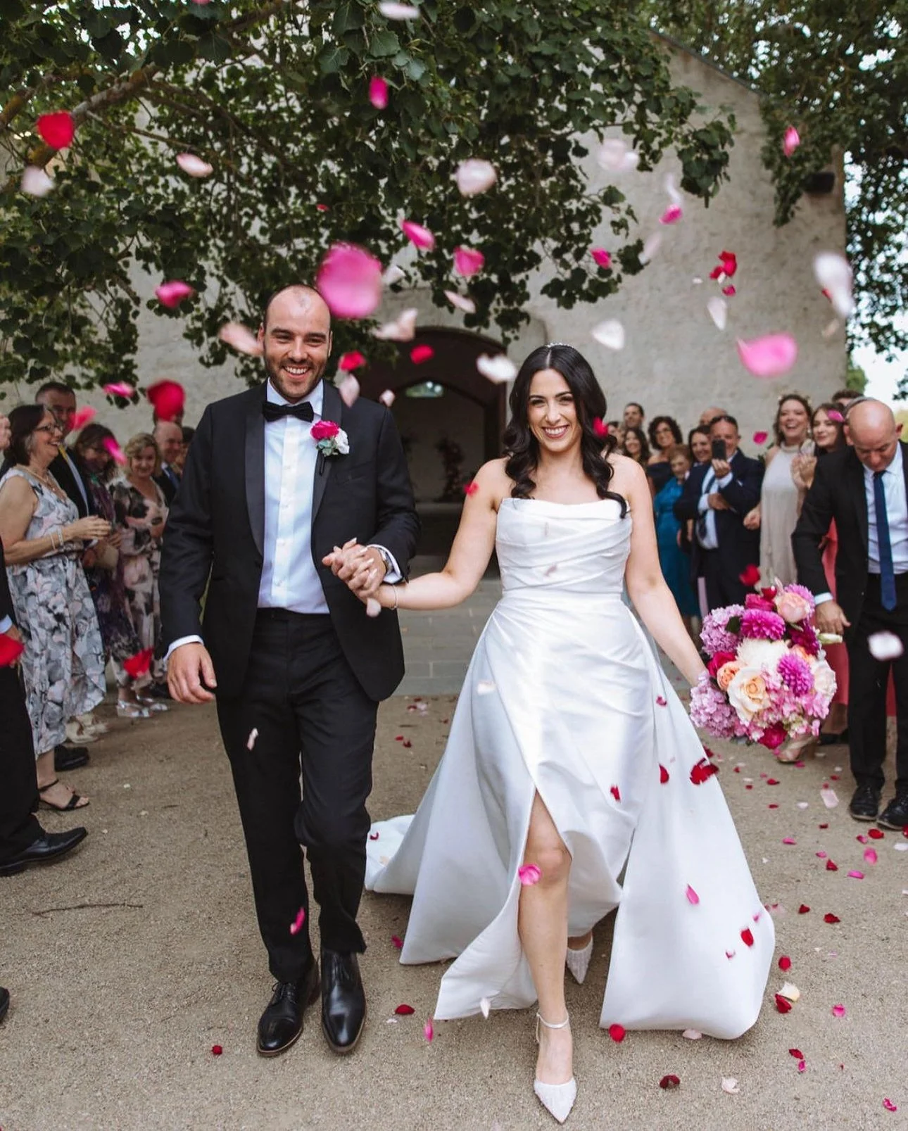 Happy bride and groom holding hands walking through falling rose petals, surrounded by smiling guests outside a stone building.