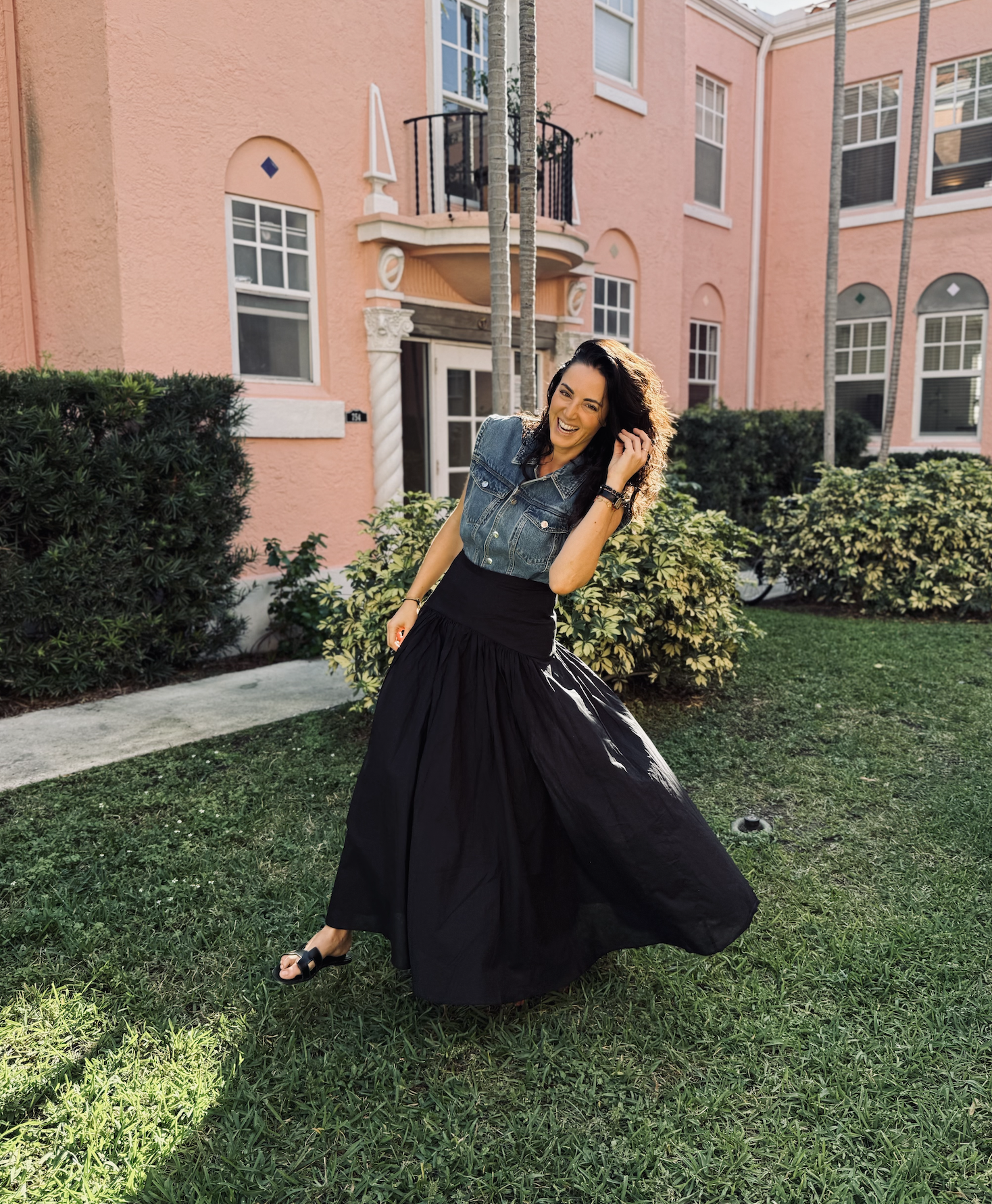 A woman in a denim vest and black long skirt is laughing while standing on a grassy lawn in front of a pink building with large windows and bushes.