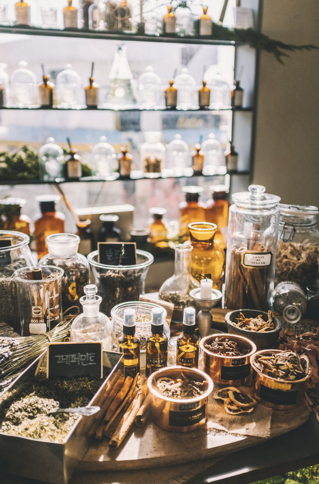 A display of various herbal remedies, oils, and dried herbs on a countertop with glass jars and bottles, set against a window with natural daylight.
