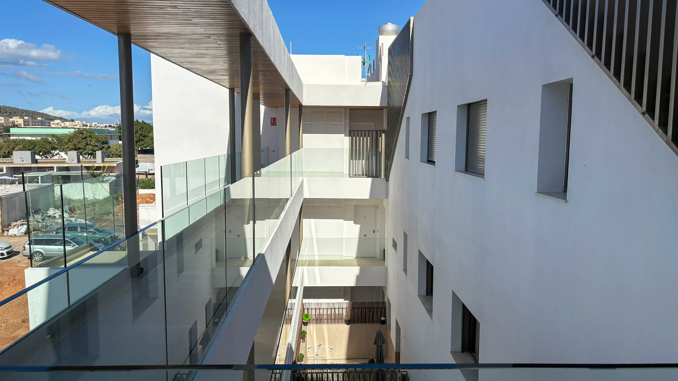 View of a modern apartment building featuring white walls, multiple windows with closed shutters, glass railings, and a courtyard below with outdoor furniture and umbrellas. Clear sky with some clouds and a parking lot with cars is visible in the bac