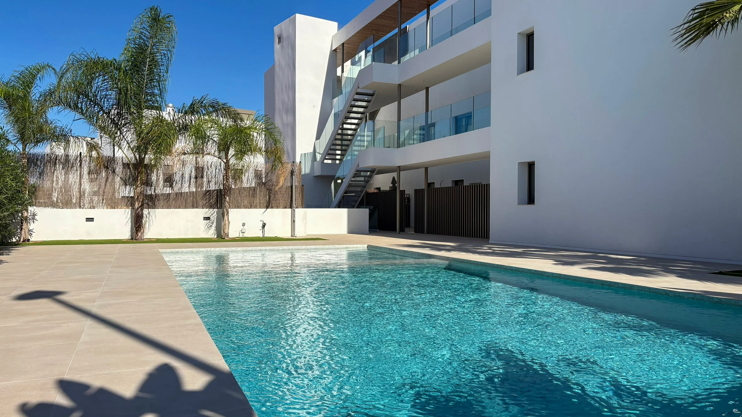 Modern multi-story apartment building with balconies and glass railings, palm trees, and a swimming pool in the foreground under a clear blue sky.