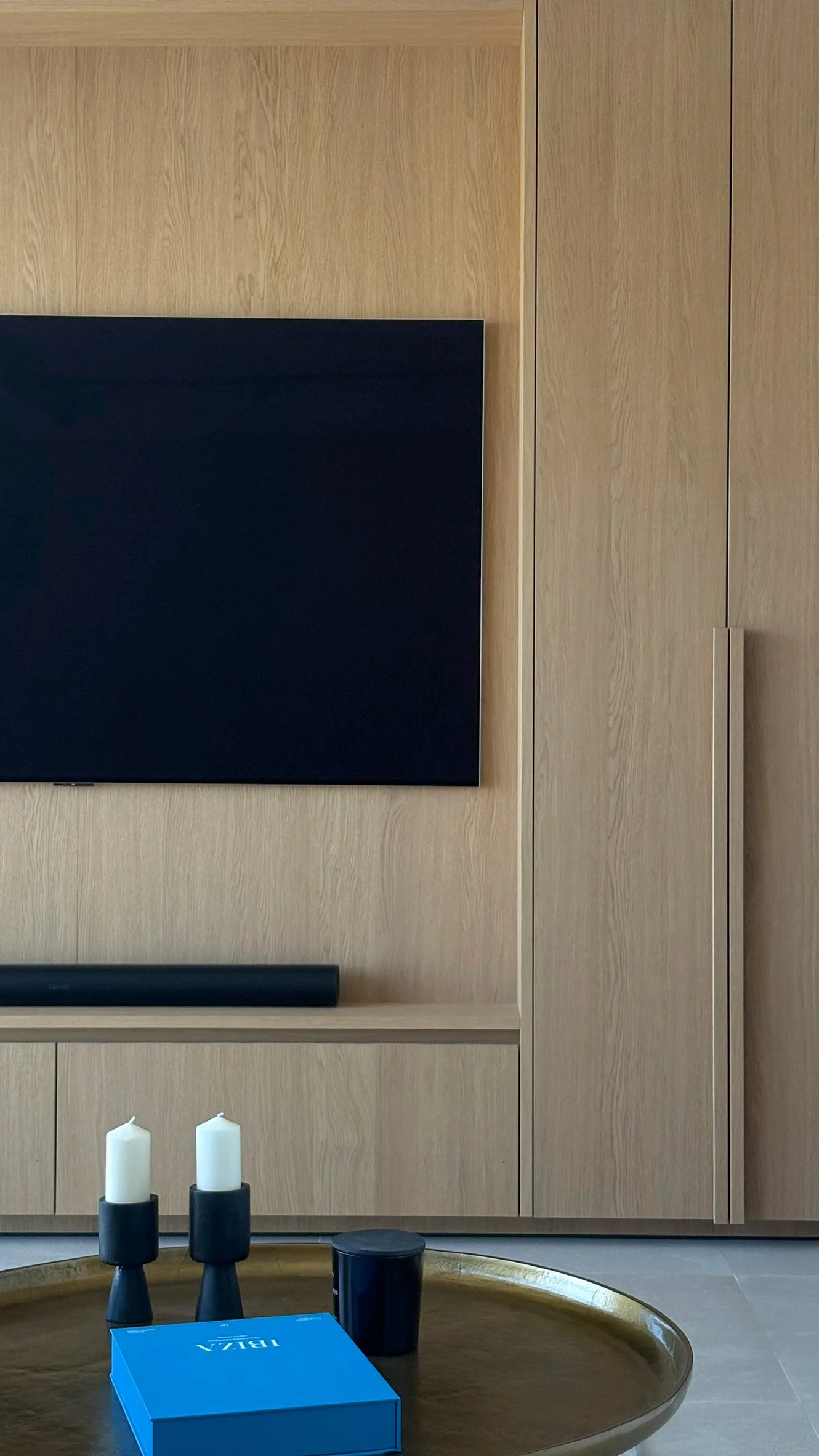 Living room with a wall-mounted flat-screen TV, light wood cabinetry, a wooden coffee table with candles and a blue box.