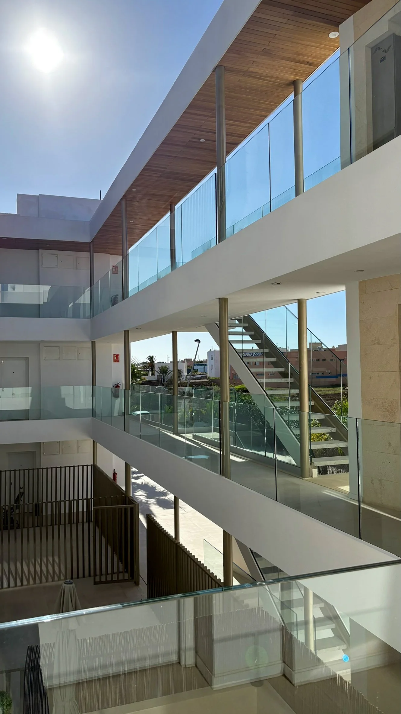 Interior view of a modern multi-story building with glass railings, stairs, and wooden ceiling panels, sunny day outside.