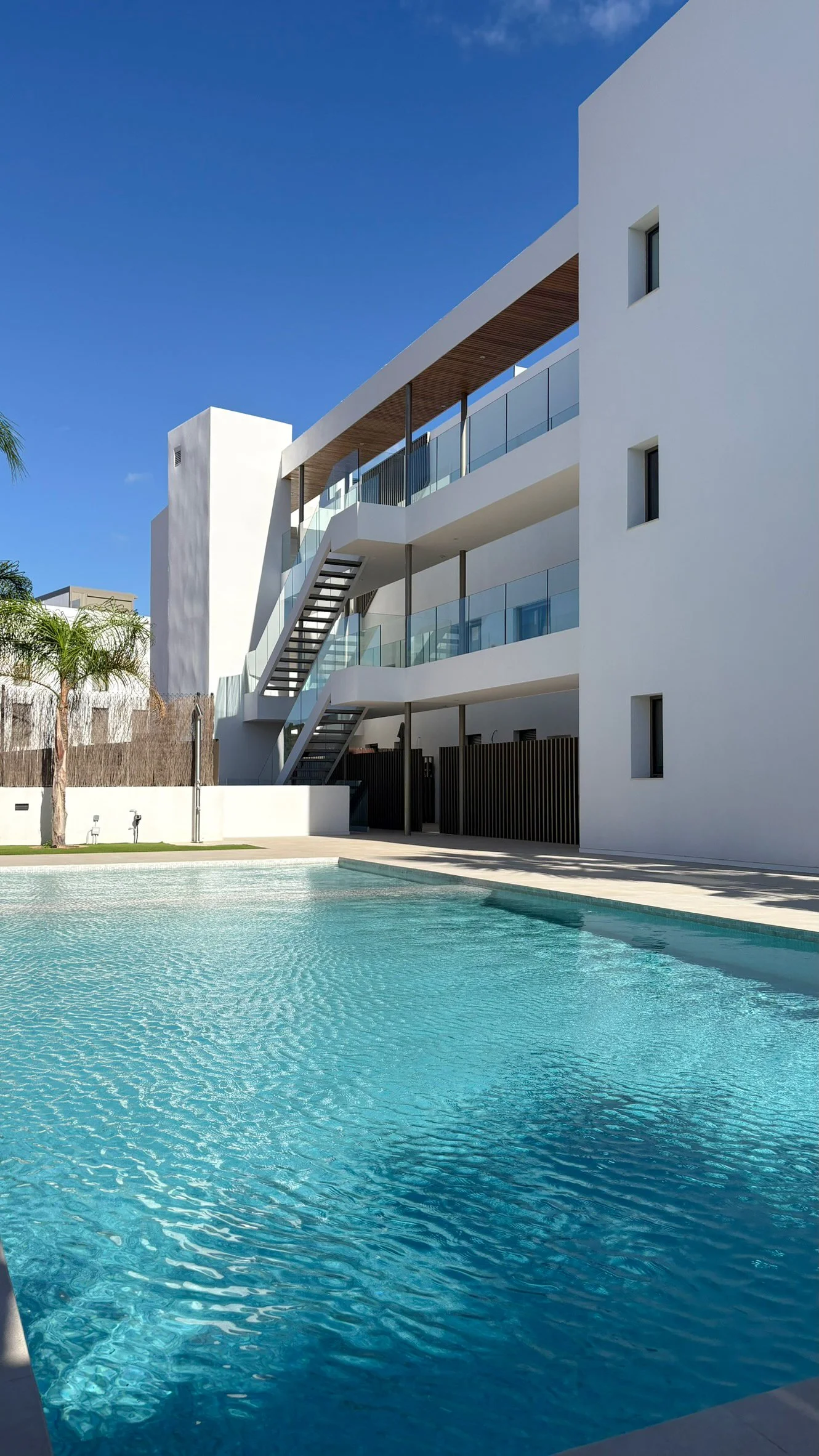 Modern white residential building with glass balconies and outdoor stairs, located next to a swimming pool under a clear blue sky.