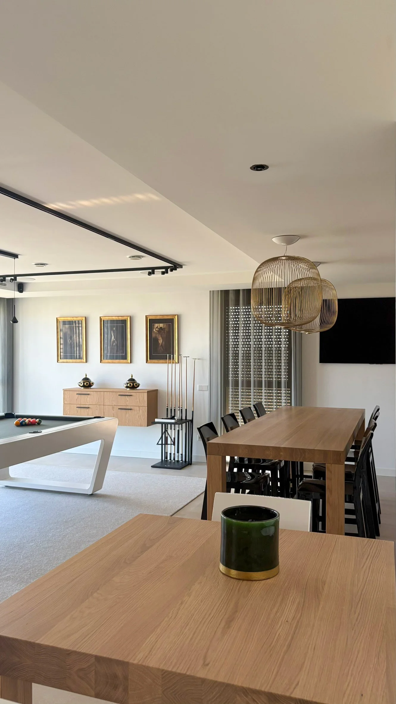 Modern living room with a pool table, wooden dining table with black chairs, and decorative pendant lights, with wall art and a window with shutters.