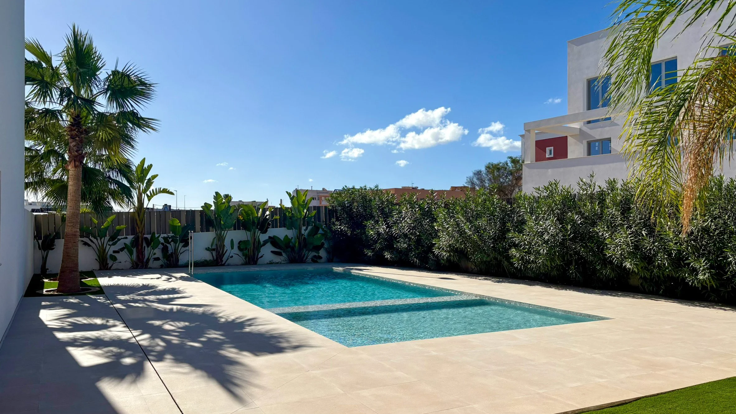 Modern backyard with a swimming pool, surrounded by palm trees, bushes, and a white residential building under a blue sky with scattered clouds.