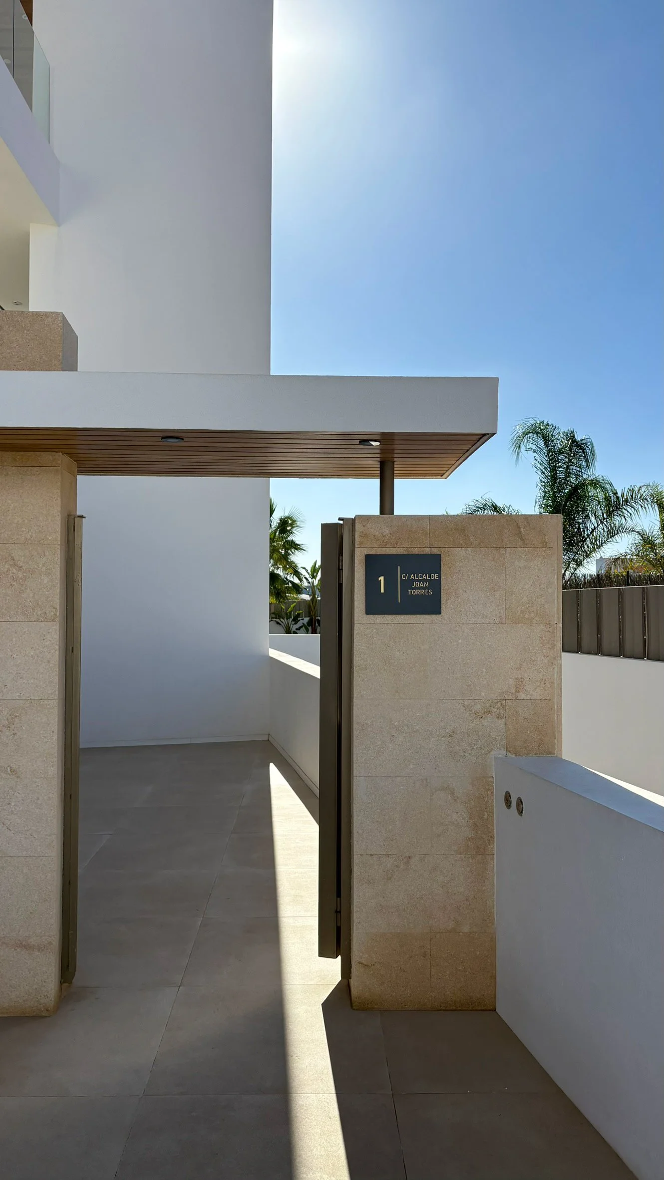 Modern building entrance with a beige stone wall, a black sign displaying address number 1 and street name in Spanish, a partial overhang, and palm trees in the background under a clear blue sky.