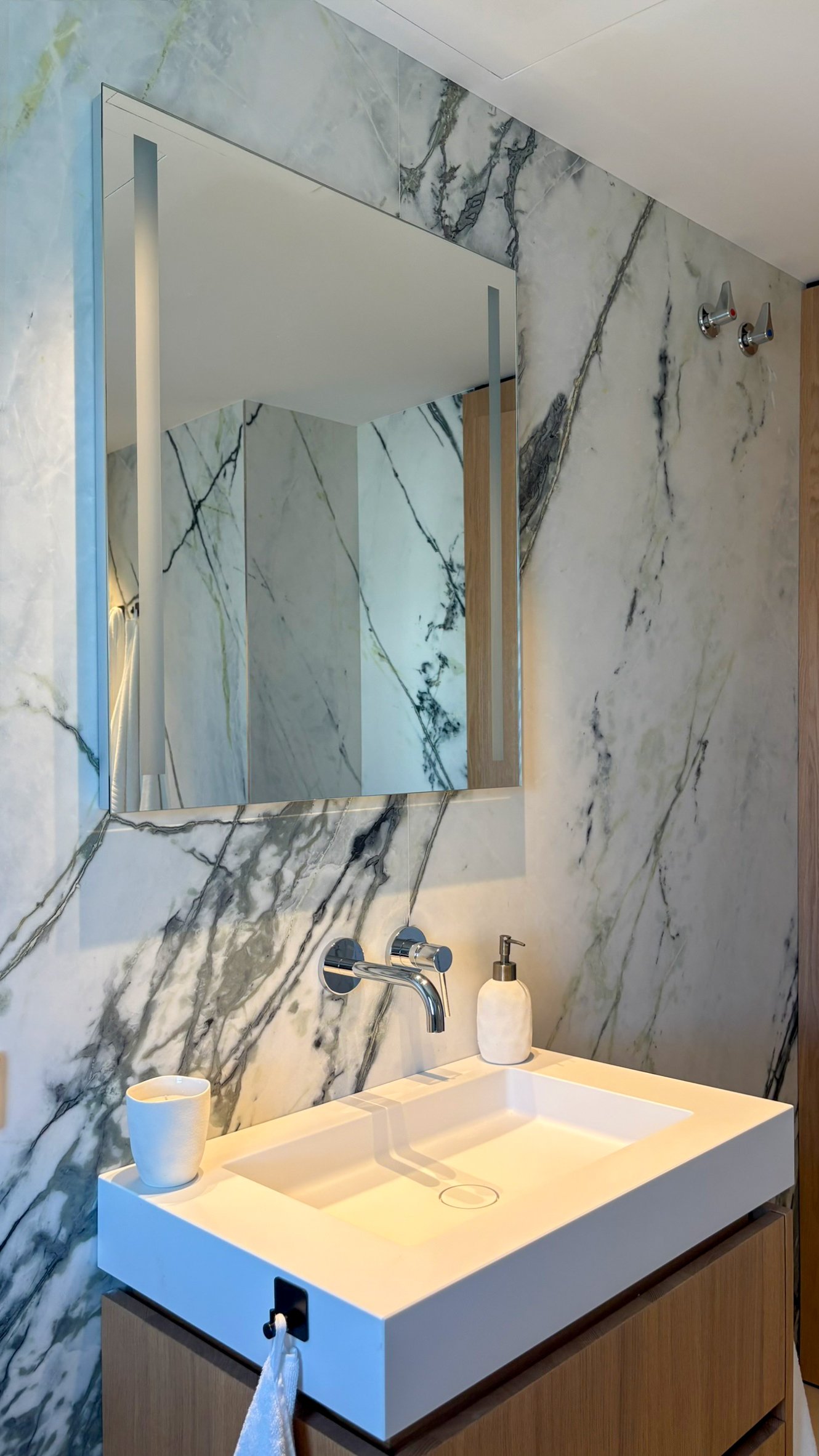 Modern bathroom with marble walls, rectangular white sink, wall-mounted faucet, mirror with built-in lighting, and wooden cabinet.