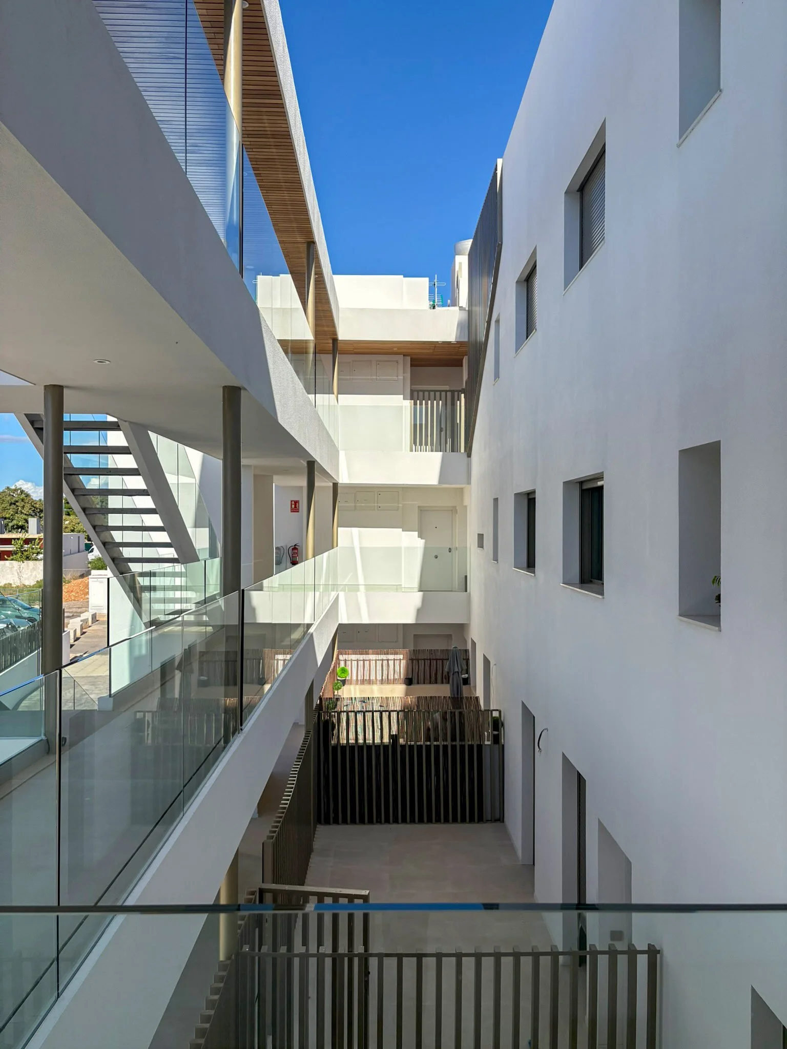 Interior view of a modern apartment building courtyard with white walls, glass balconies, metal staircases, and multiple small rectangular windows, under a clear blue sky.