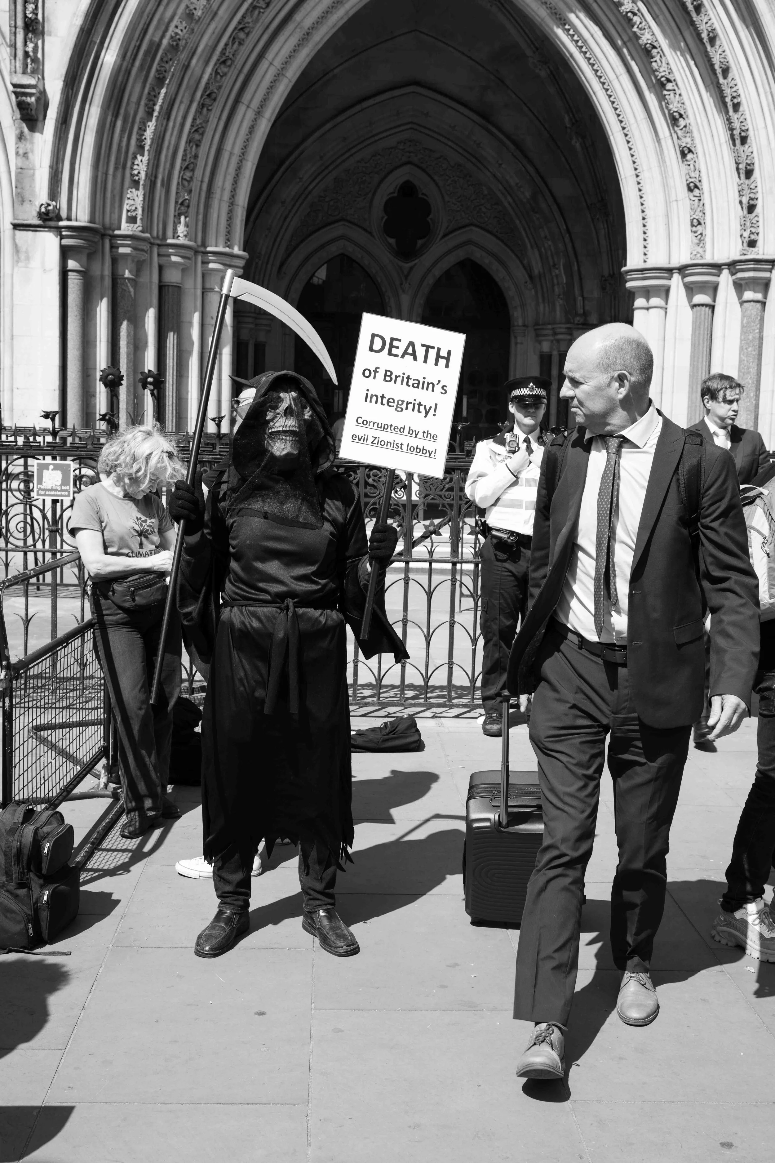 A man is exiting The Royal Courts of Justice and looks back at a Pro Palestinian demonstrator. The demonstrator is dressed in black with a mask and is holding a scythe and a sign saying 'Death of British Integrity'