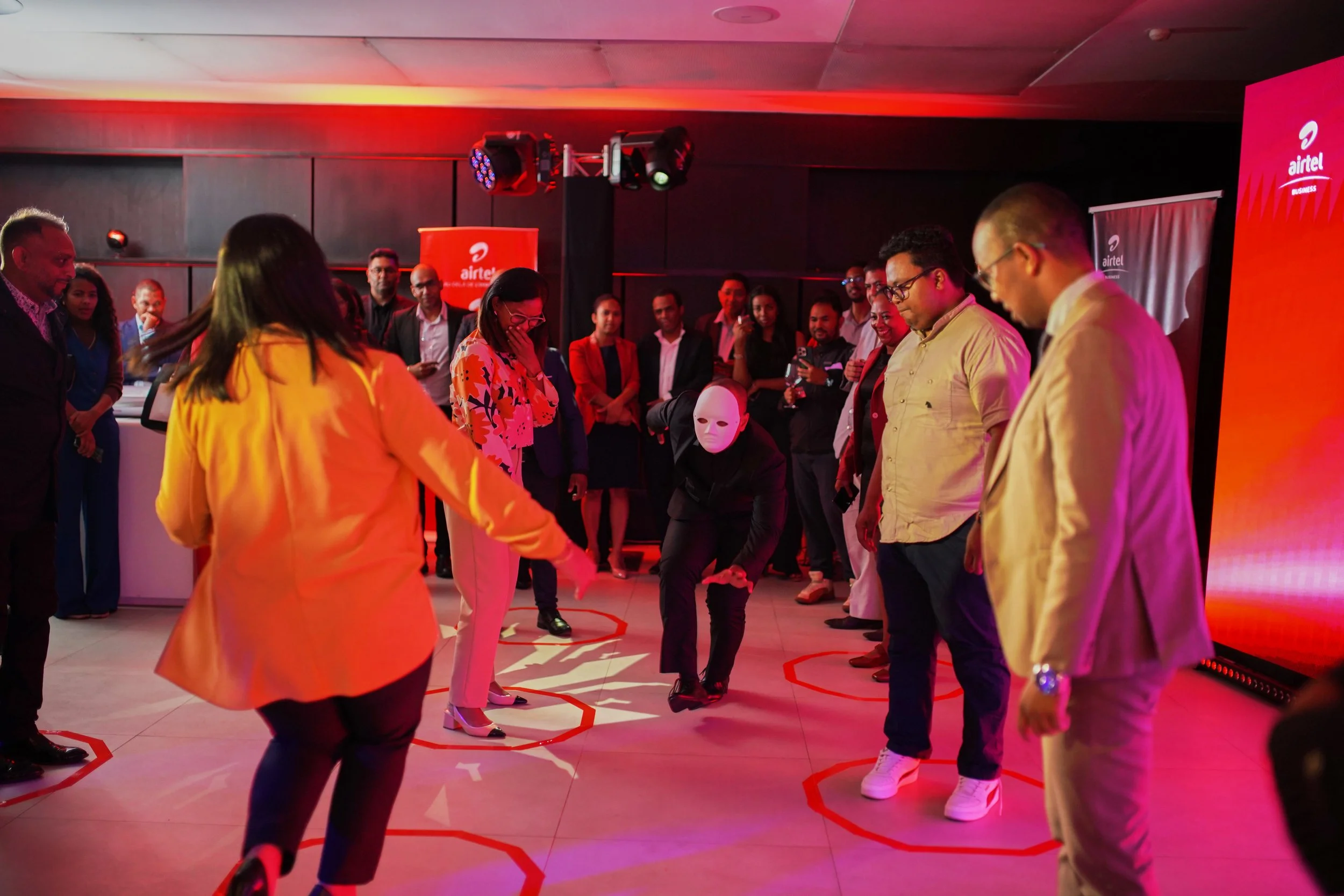 Group of people participating in a team-building activity with one person wearing a white mask, inside a room with red lighting and branding for Airtel.