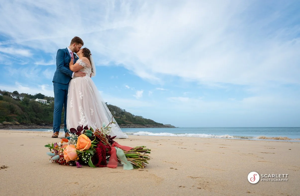 A bride and groom sharing a kiss on the beach with a bouquet of flowers in the foreground.