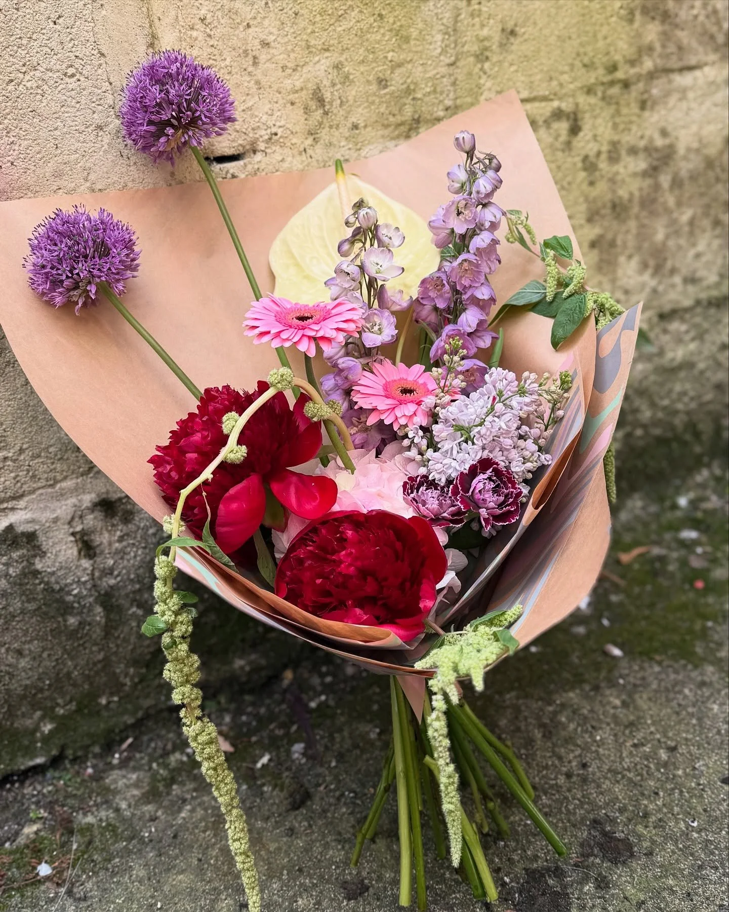 A colorful bouquet of various flowers wrapped in pink paper, placed against a concrete wall.