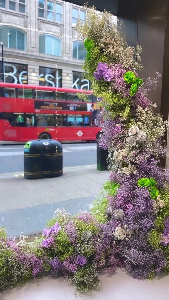 A large scale floral arrangement with purple, white, and green flowers by a window, with a city street scene including a double-decker bus and a building with large windows in the background.