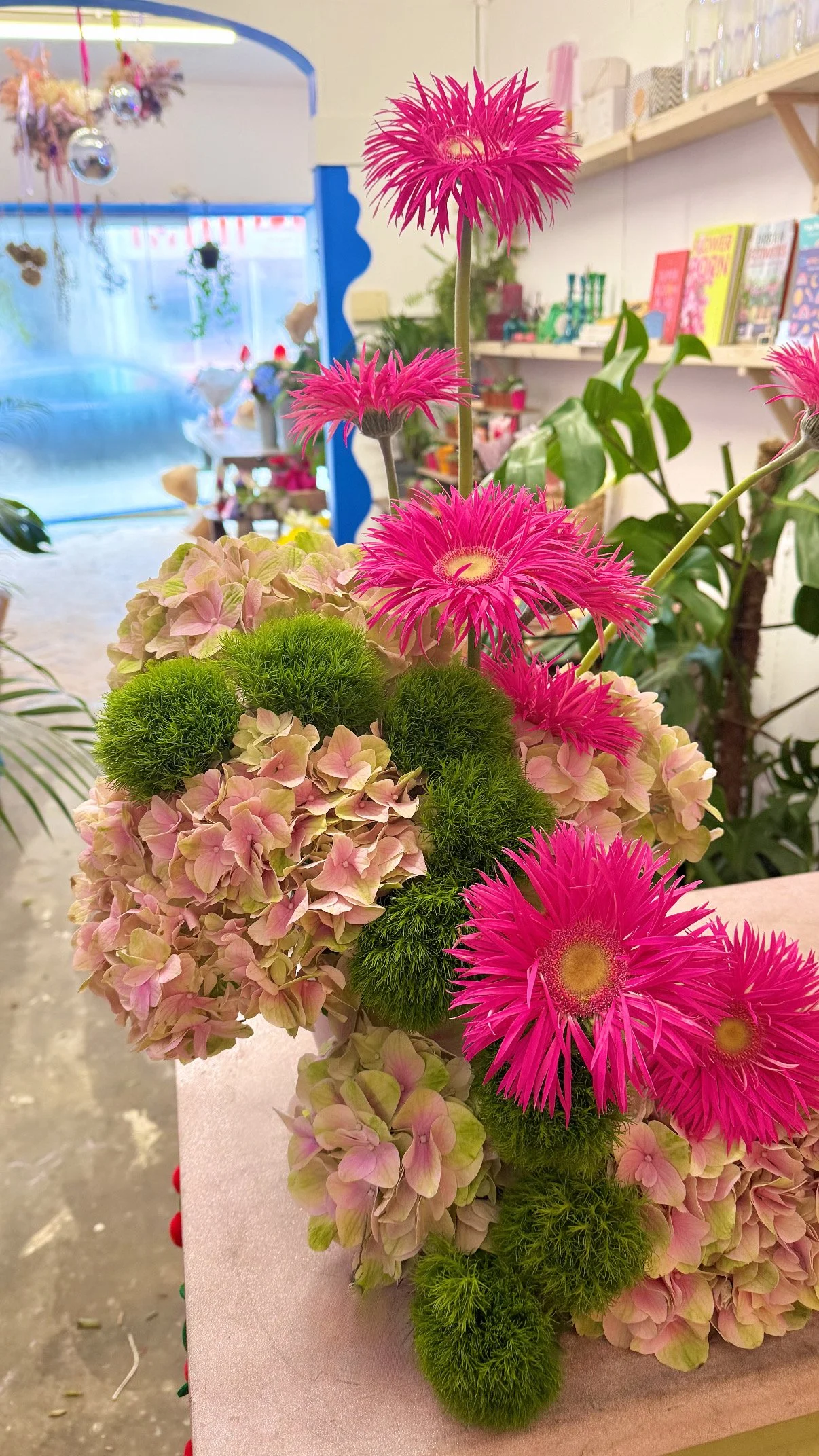 A colorful floral arrangement featuring bright pink spider gerbera, pale pink hydrangeas, and green fluffy dianthus balls on a pink surface inside a flower shop.