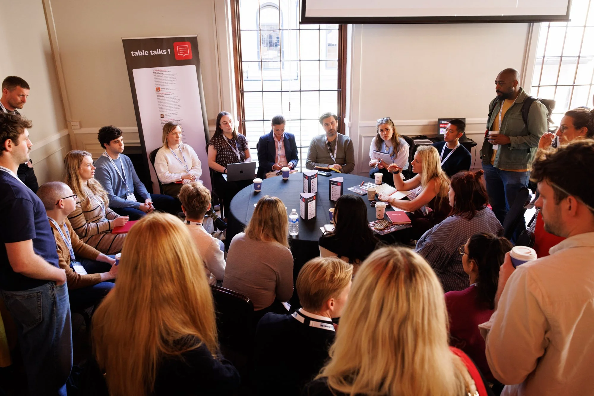 A diverse group of people seated around a round table during a meeting or workshop in a well-lit room with large windows. Some individuals are taking notes or holding cups, and one person is standing and speaking.