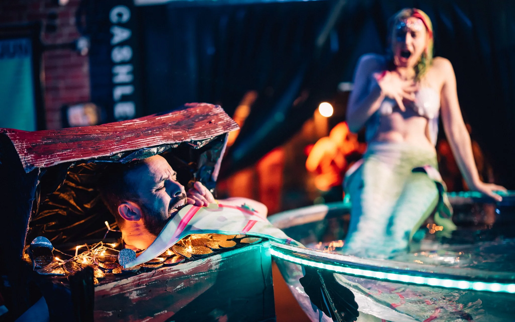  man dressed as treasure chest bites mermaids tail at Party.