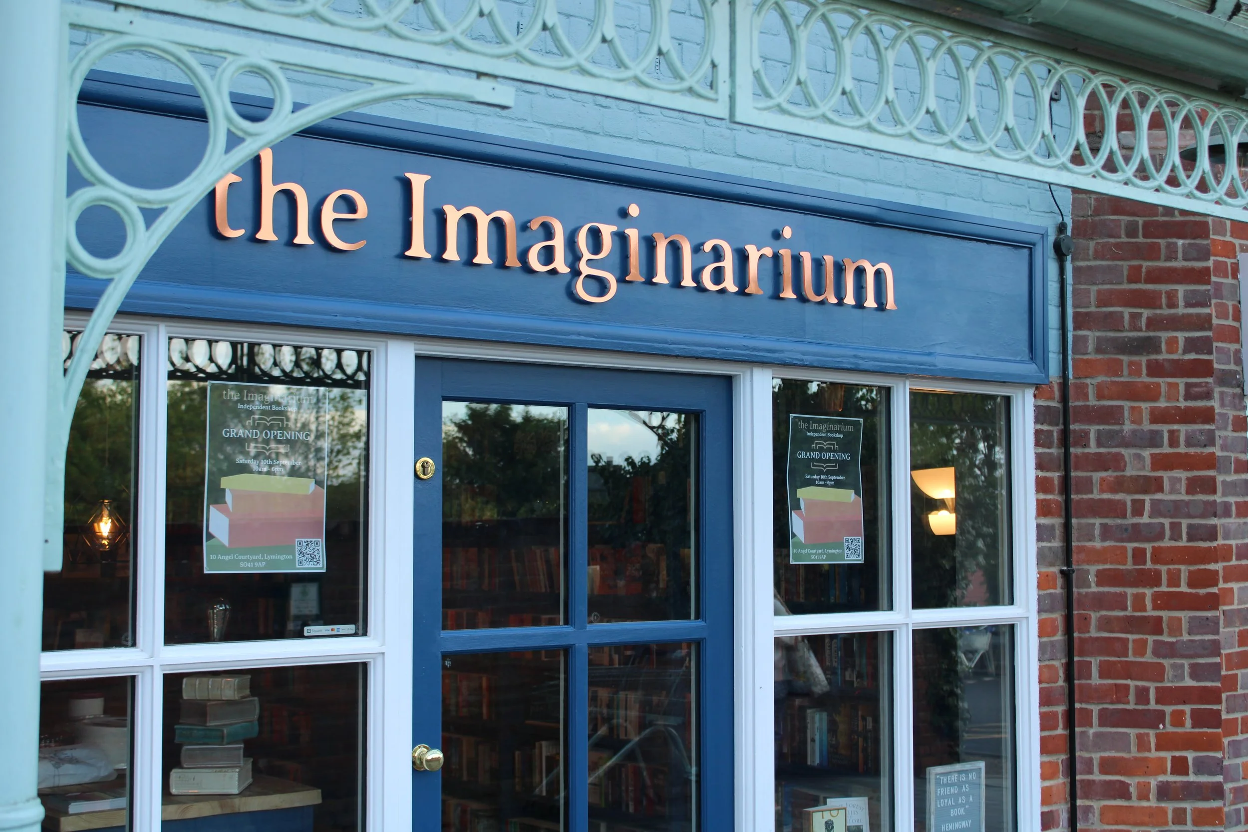 The storefront of a bookstore named 'the Imaginarium' with a blue sign, glass windows displaying signs about a grand opening, and the interior visible through the windows includes bookshelves and books.