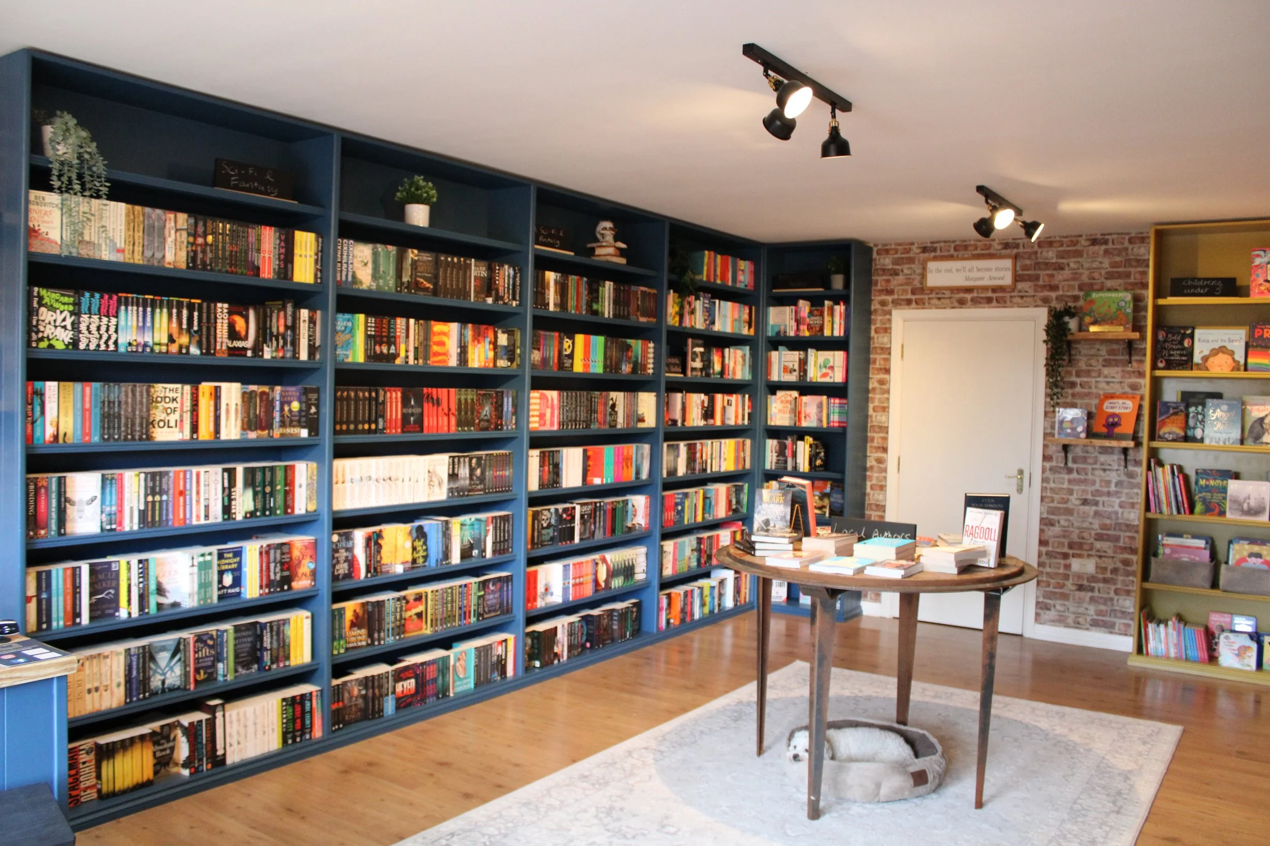 A cozy bookstore with blue bookshelves filled with books, a round wooden table with stacks of books, and a small dog resting in a pet bed underneath the table. The room has a brick wall, hardwood floors, and ceiling track lighting.
