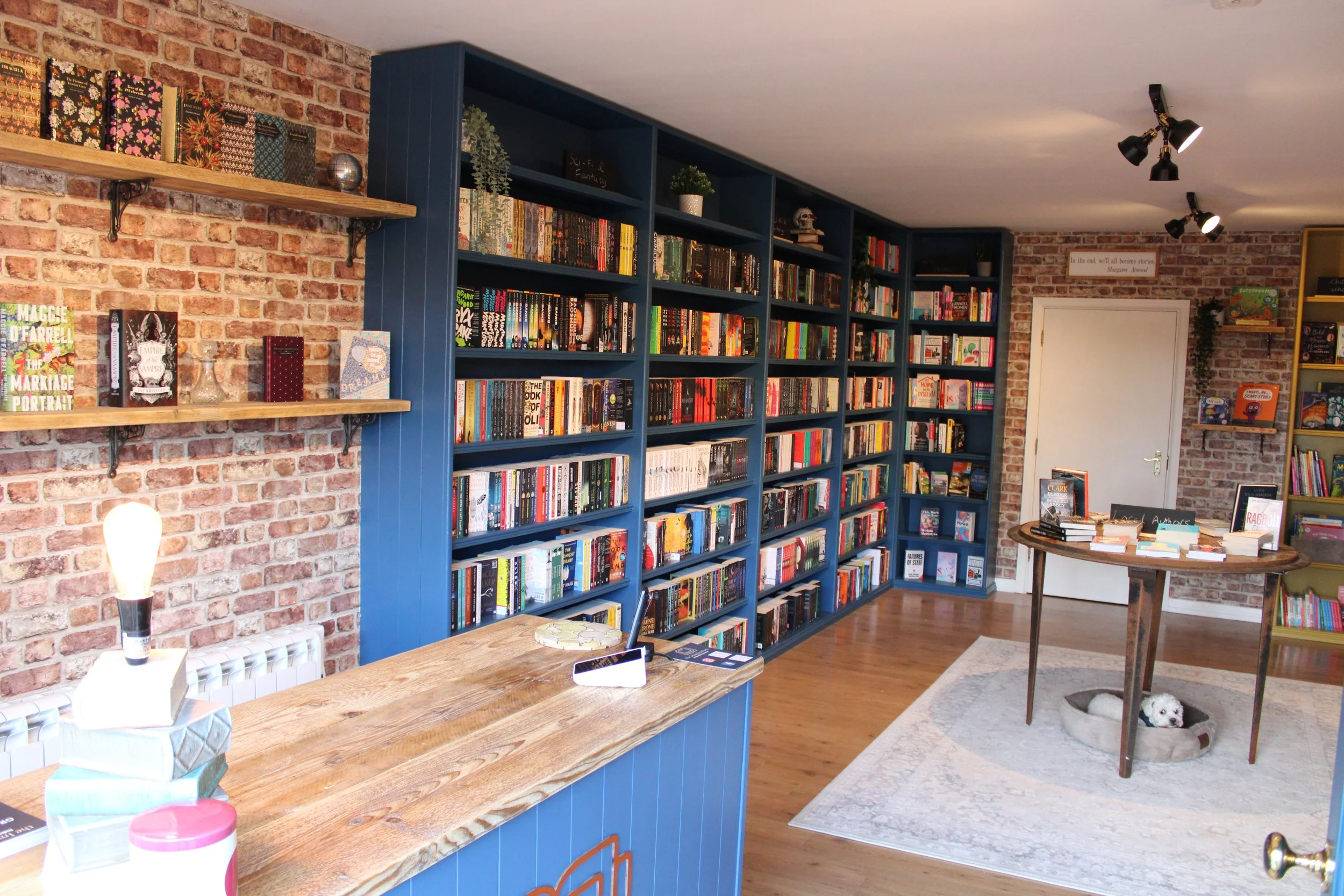 Interior of a bookstore with blue bookshelves filled with books, brick walls, and a wooden counter in the foreground. There is a small round table with books and a dog resting in a pet bed underneath it, placed on a white rug.