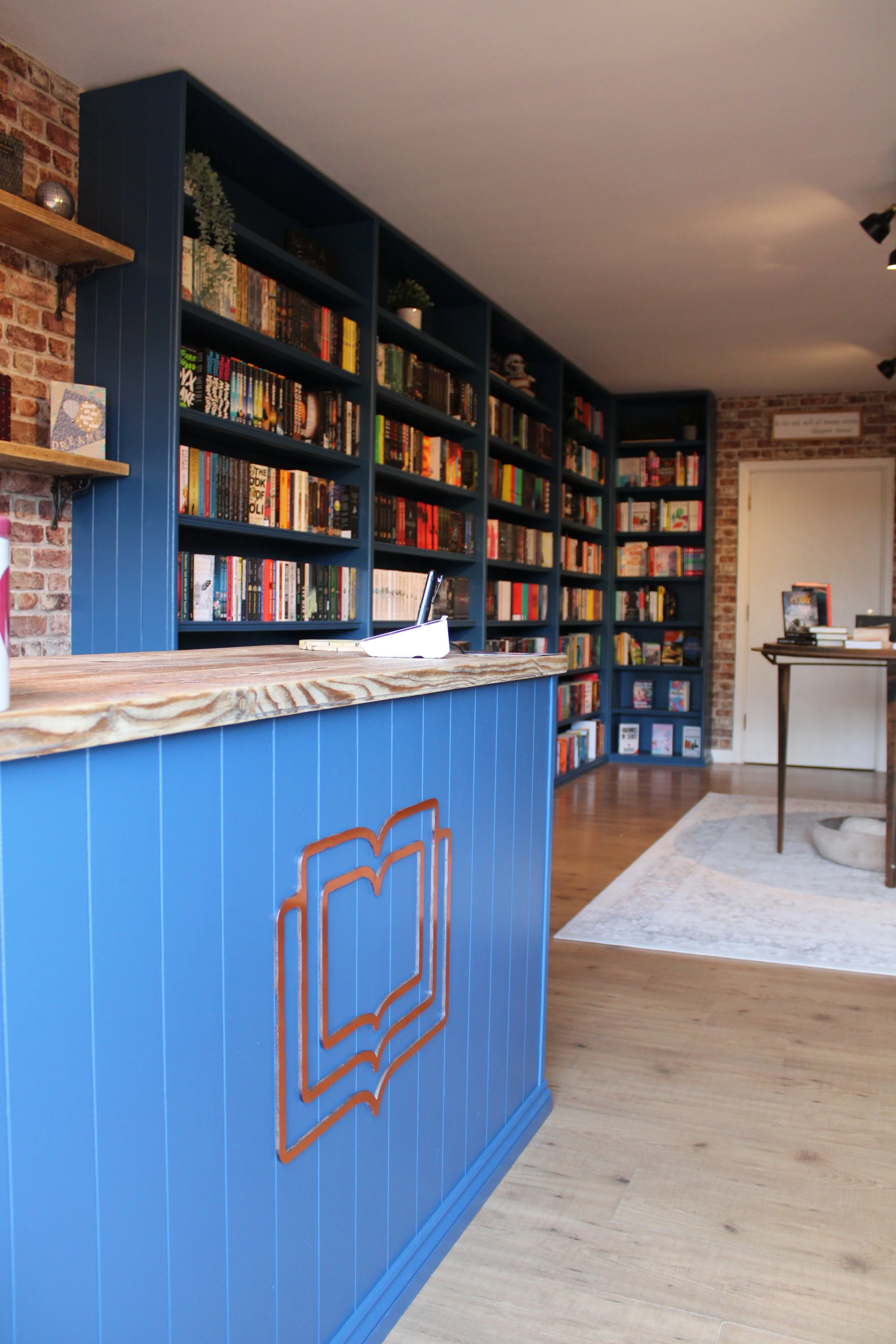 Interior of a cozy bookstore or library with blue wooden shelves filled with books, a brick wall, and wooden flooring.
