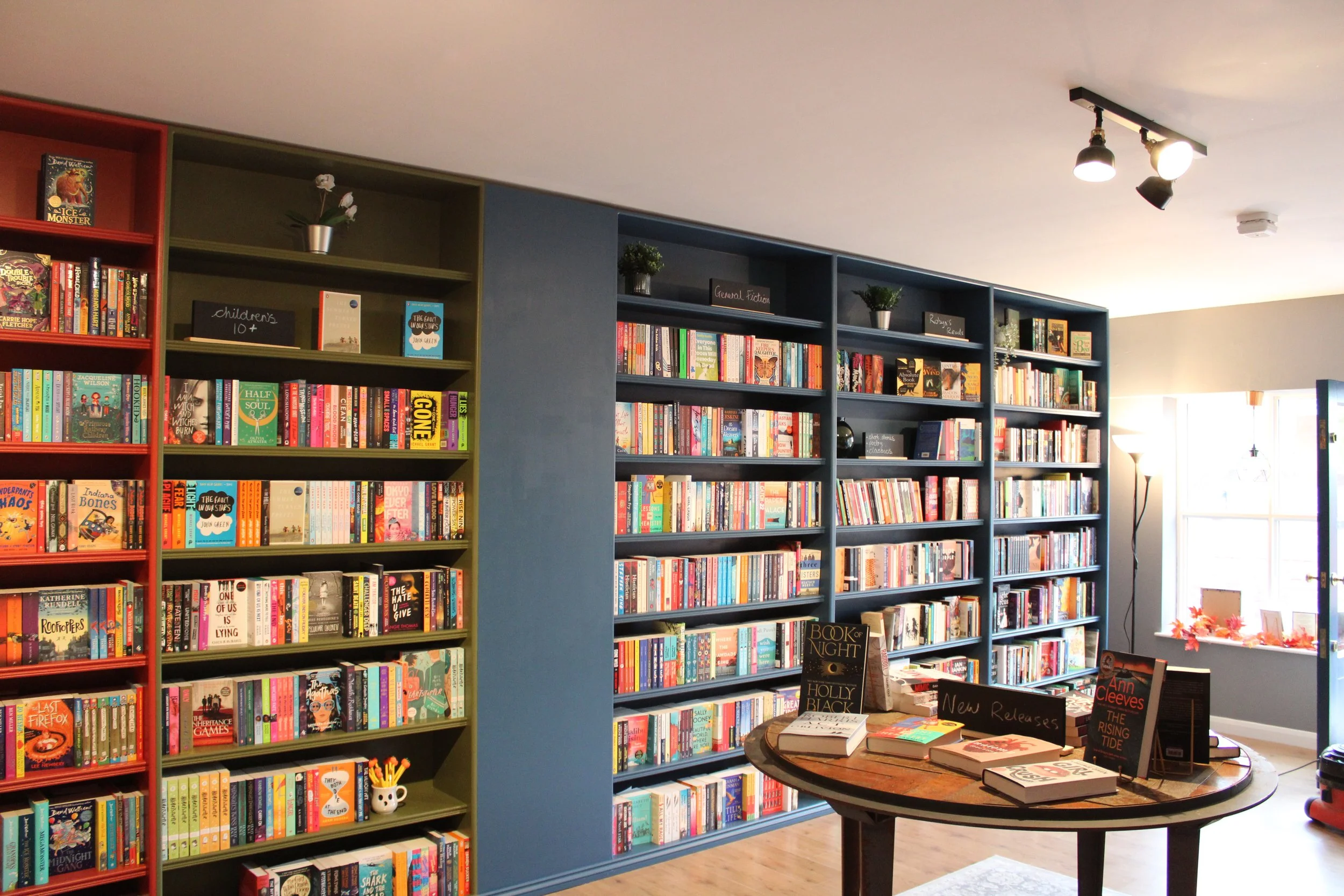 Bookshelves filled with a variety of books in a bookstore or library, with a round table displaying new releases and signs indicating book sections.