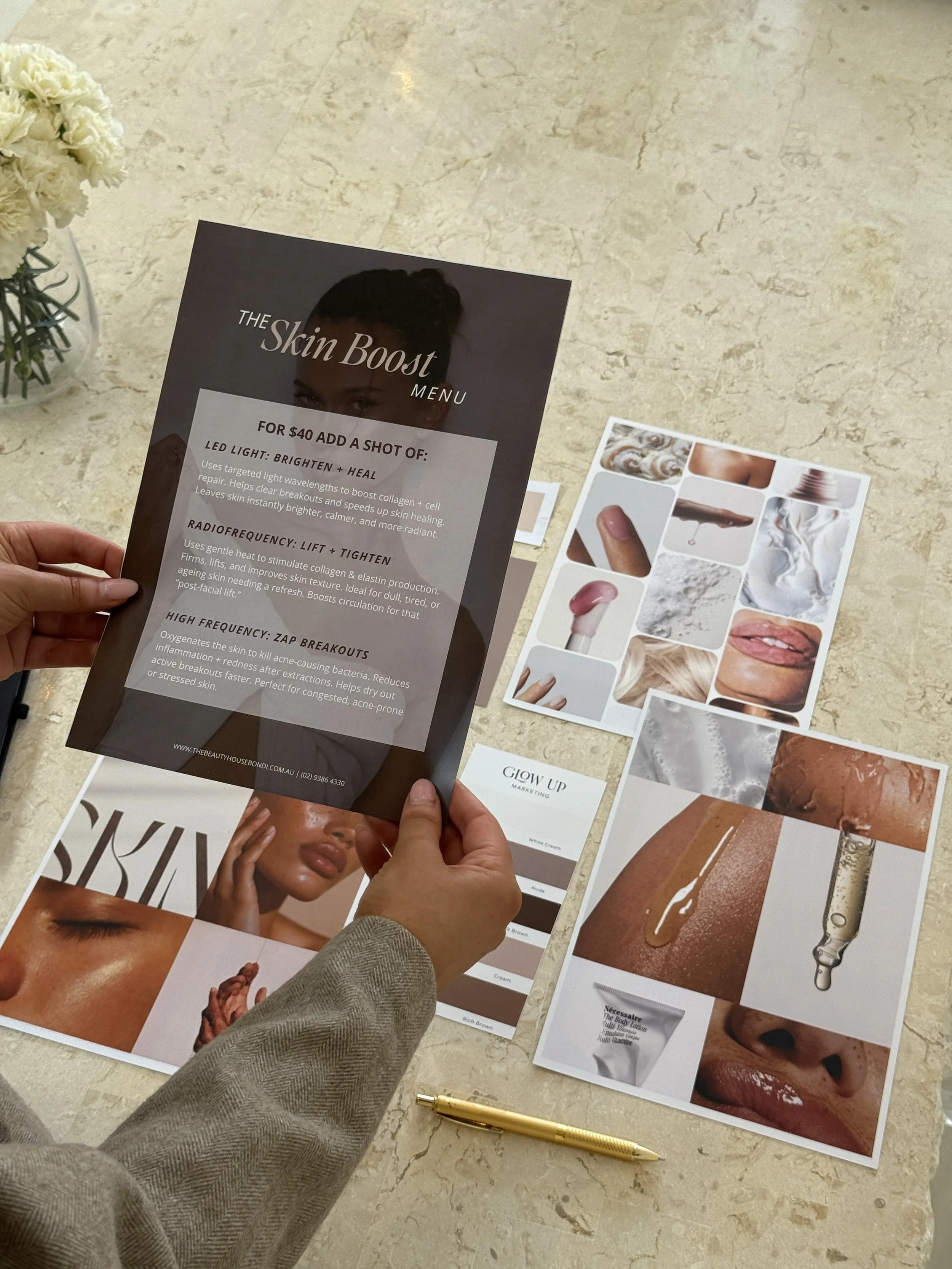 Person holding a black promotional flyer for a skincare menu titled 'The Skin Boost Menu,' with three sheets of skincare and beauty product images and a gold pen on a beige marble table, near a small bouquet of white flowers.