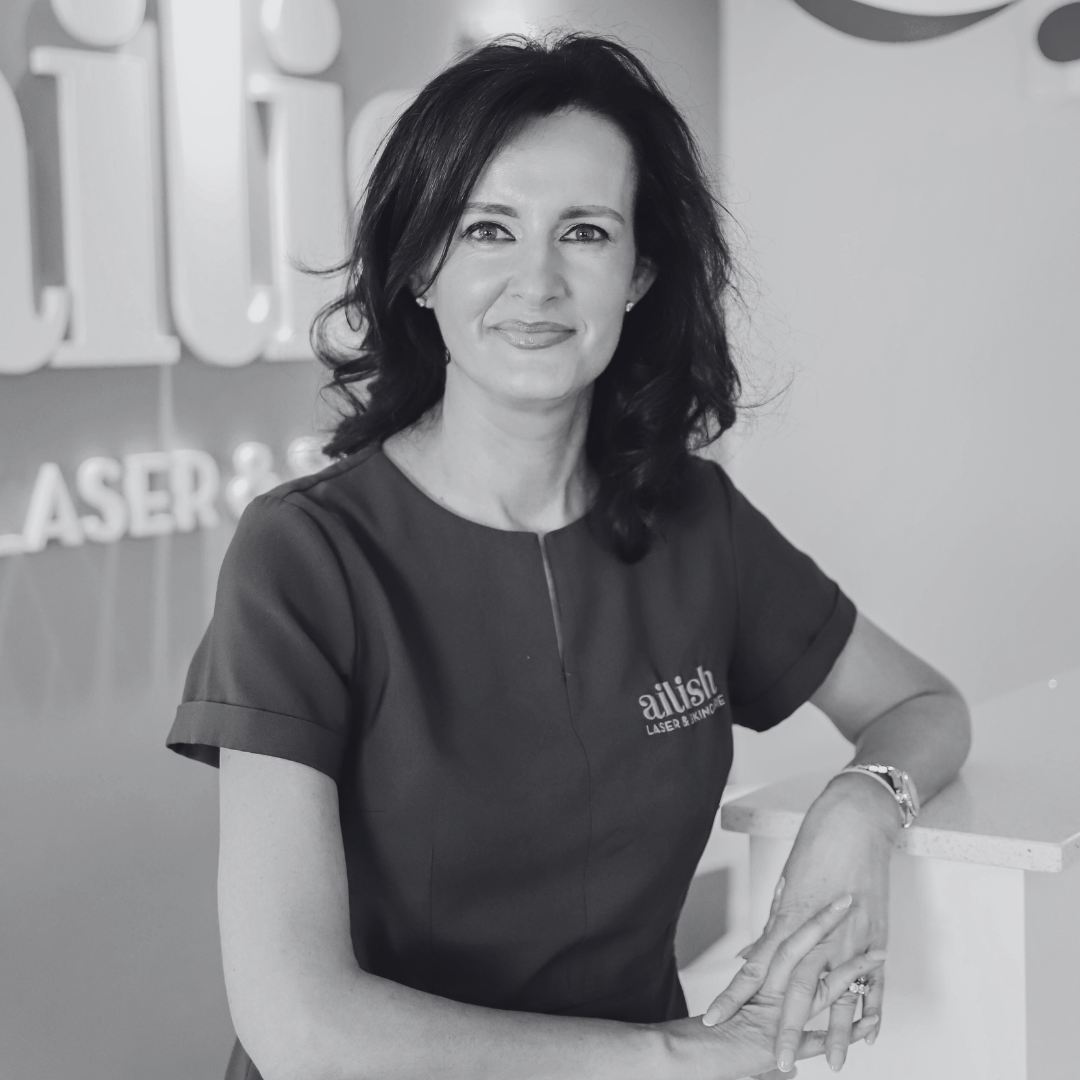 A woman with dark, wavy hair smiling, wearing a dark top with the logo 'ailish' and 'Laser & Skin Care', sitting at a reception counter in a modern office or clinic setting.