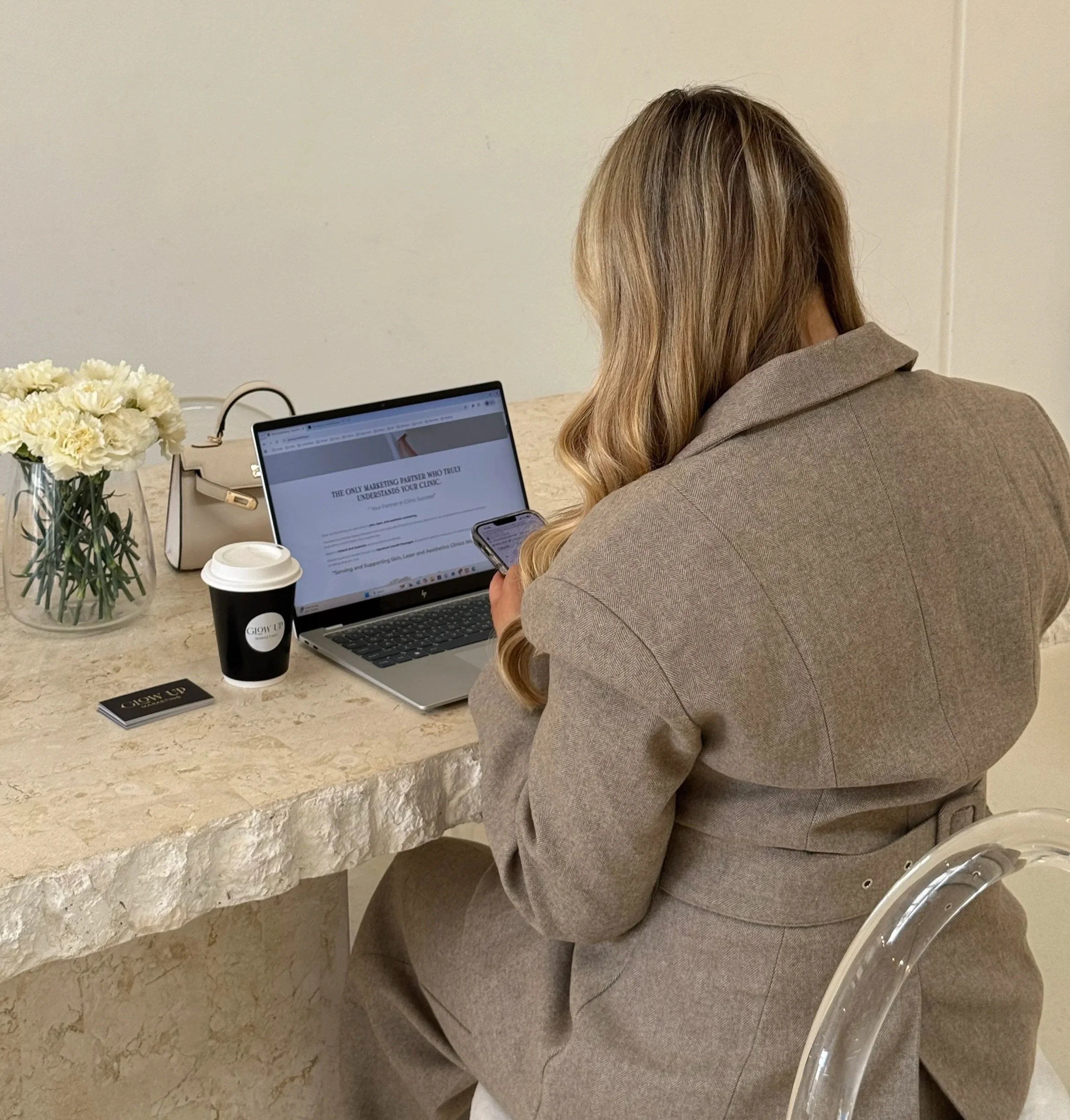 A woman with long blonde hair working on a laptop and looking at her smartphone at a marble table. The table has a vase of white flowers, a coffee cup, a handbag, and business cards.