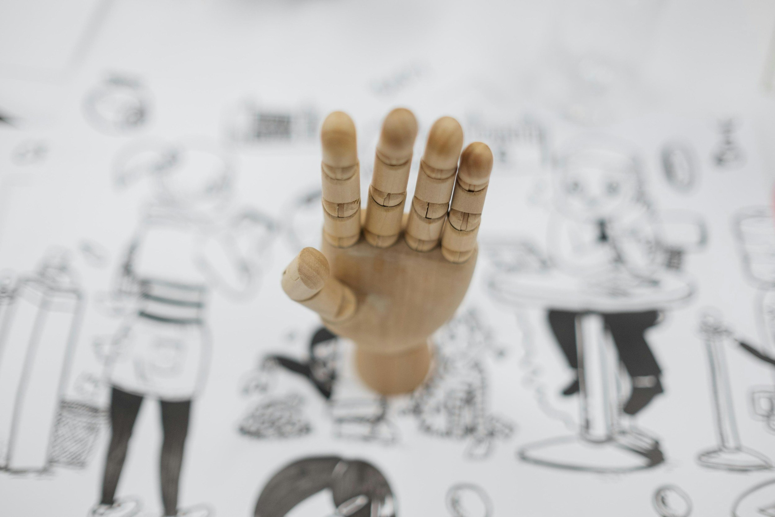 A wooden artist mannequin hand with fingers slightly bent, against a blurred background with black and white illustrations of people.