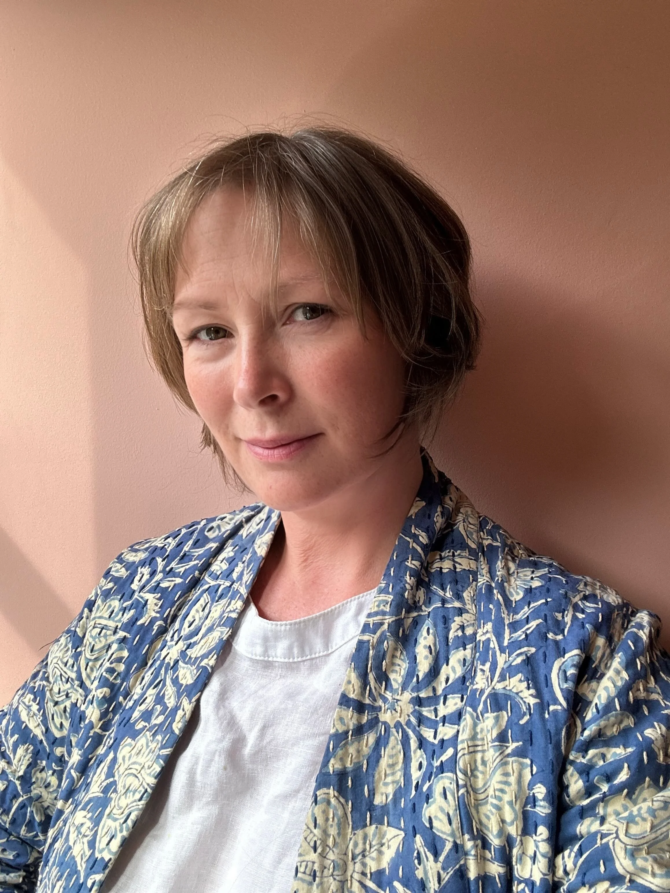 A woman with short brown hair and fair skin, smiling softly, wearing a blue and white patterned jacket over a white shirt, standing against a pink wall.