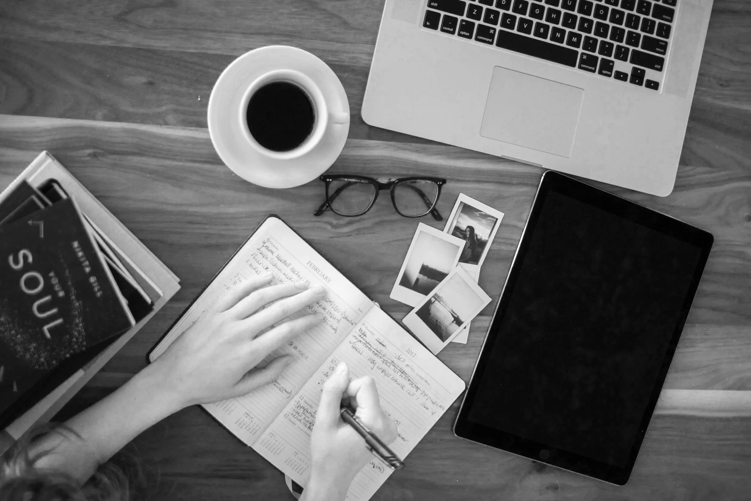 Overhead view of a workspace with a laptop, tablet, glasses, photos, a coffee cup, an open planner, and a person writing in the planner on a wooden table.