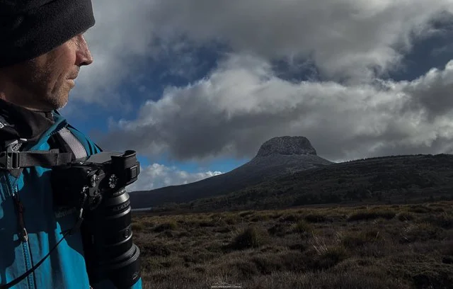 A person wearing a blue jacket and a black hat holding a camera, standing in a landscape with a mountain and cloudy sky.