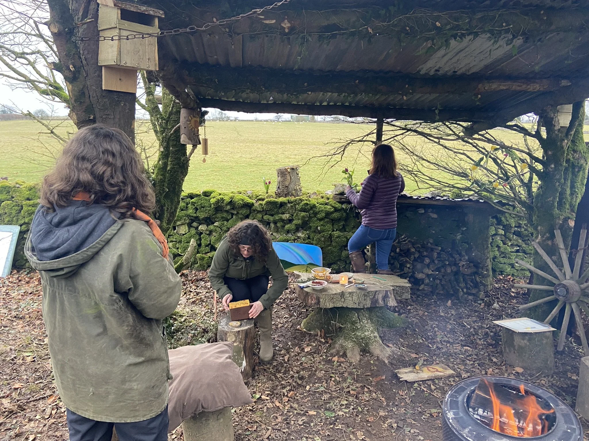 Three women are in an outdoor setting with moss-covered rocks and trees. One woman is standing with her back to the camera, looking at other two women. One woman is seated on a tree stump, holding a small wooden object, while another woman is standin