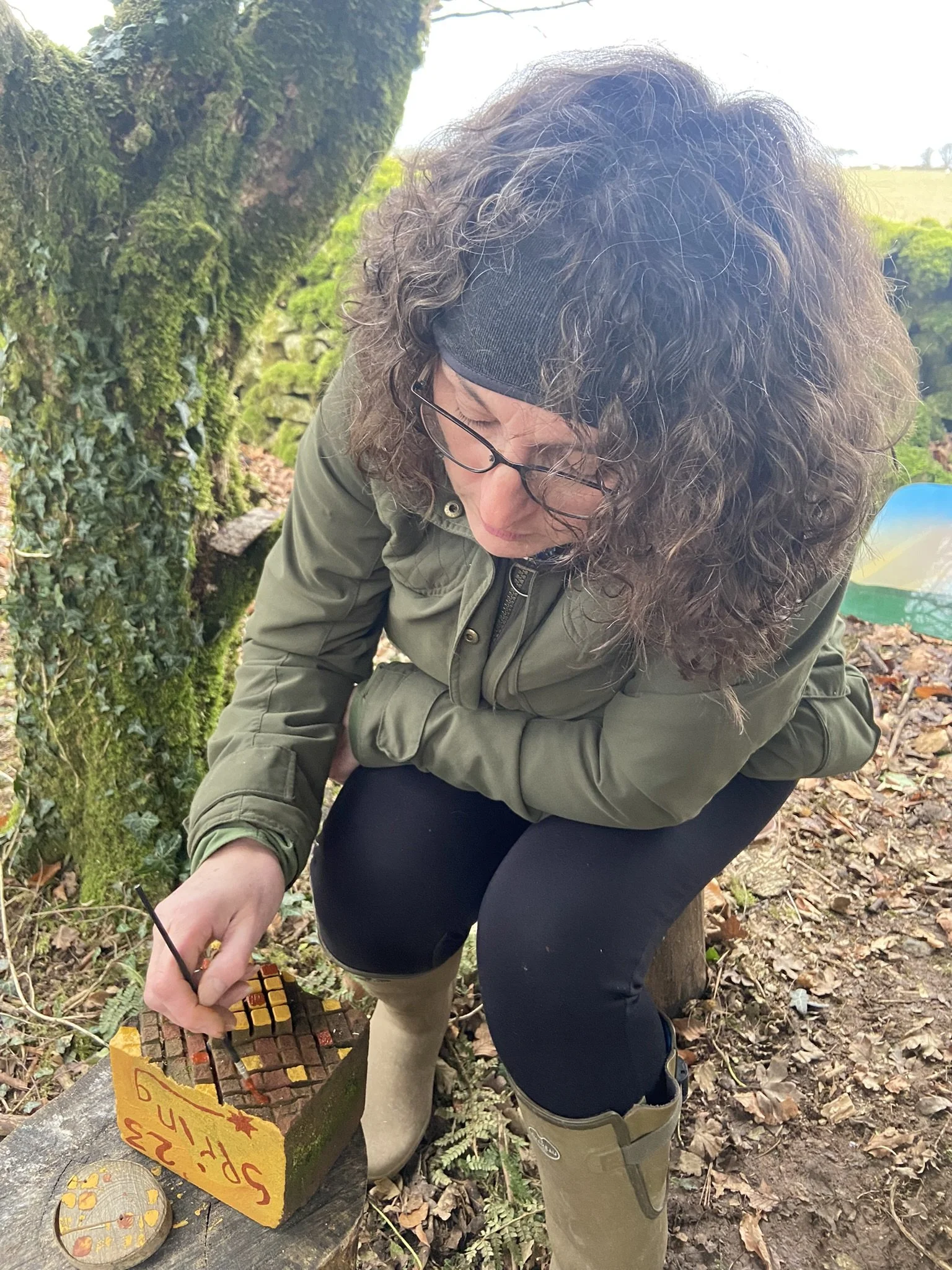 Woman with curly hair, glasses, and a black beanie, kneeling outdoors, using a small chisel to carve or inscribe a wooden block that has a yellow sticker with red writing on it.