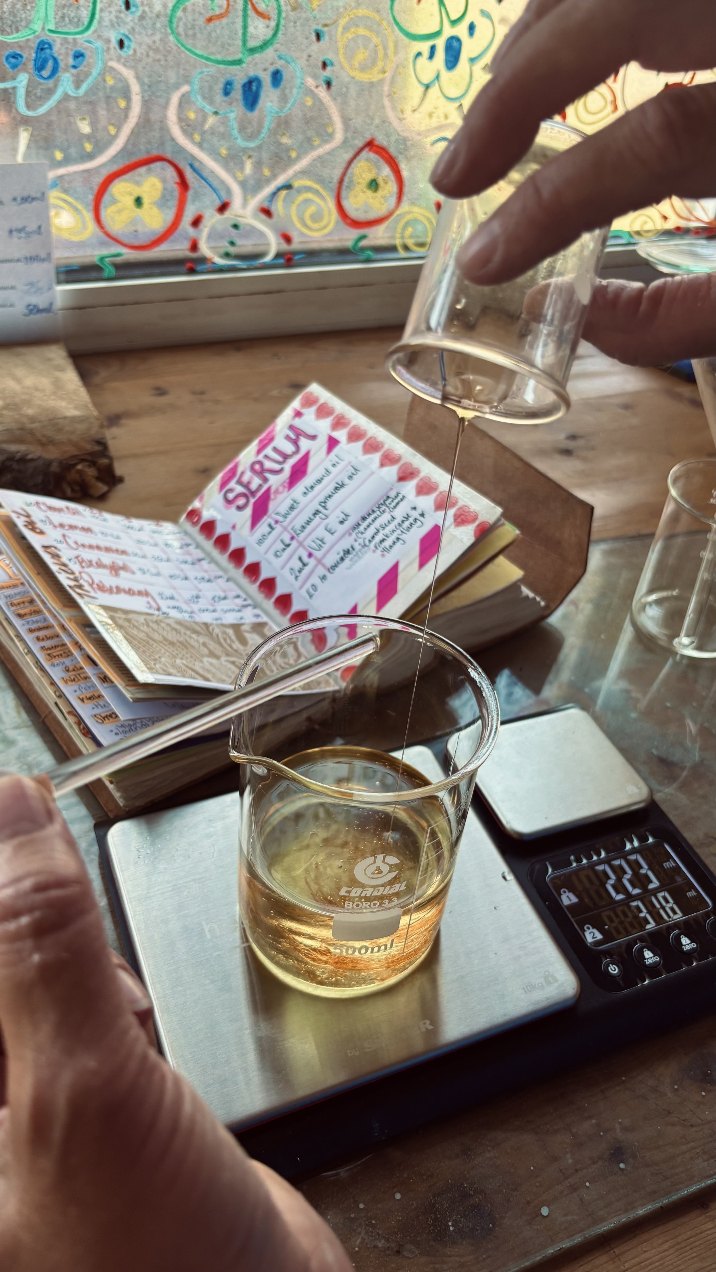 Lisa Jo pouring a clear liquid into a beaker on a digital scale, with notebooks and a colourful decoration in the background.