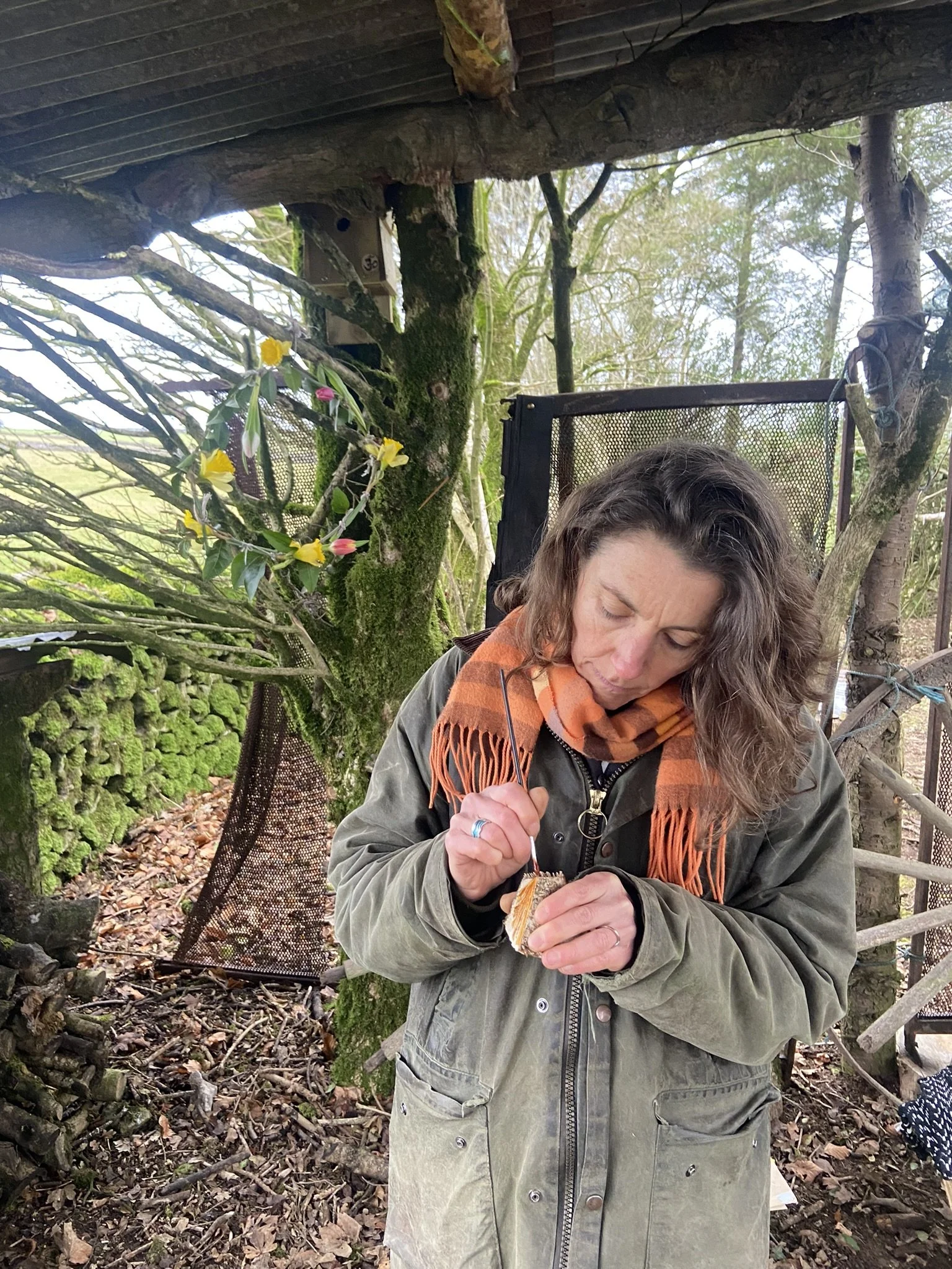 A woman with wavy brown hair wearing a green jacket and orange scarf, holding a butterfly and a fork, in an outdoor wooded area.
