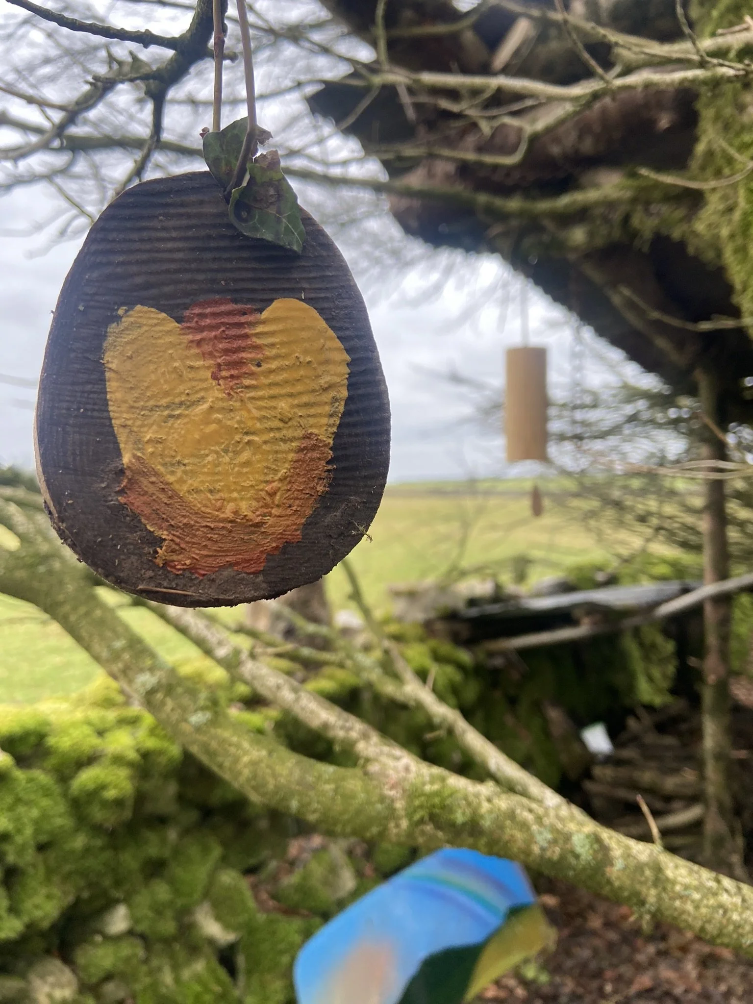 A painted wooden slice with a yellow heart and a red nipple, hanging from a tree branch with a string outdoors. In the background, there is a blurred blue object and a wind chime hanging from another branch.
