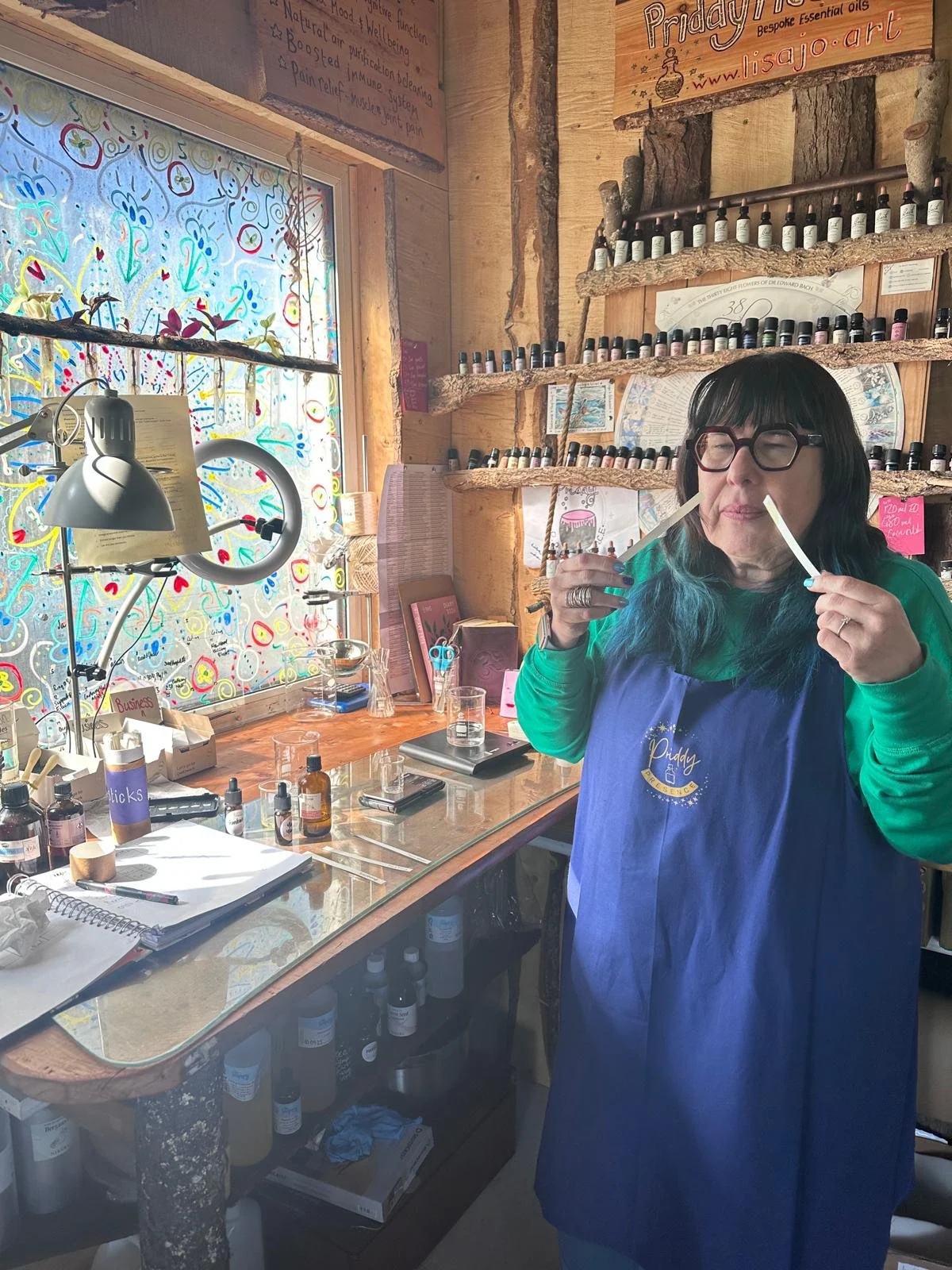 Woman with glasses and teal hair testing essential oils in a rustic studio with shelves of small bottles, a colorful window, and a workbench with notebooks and bottles.