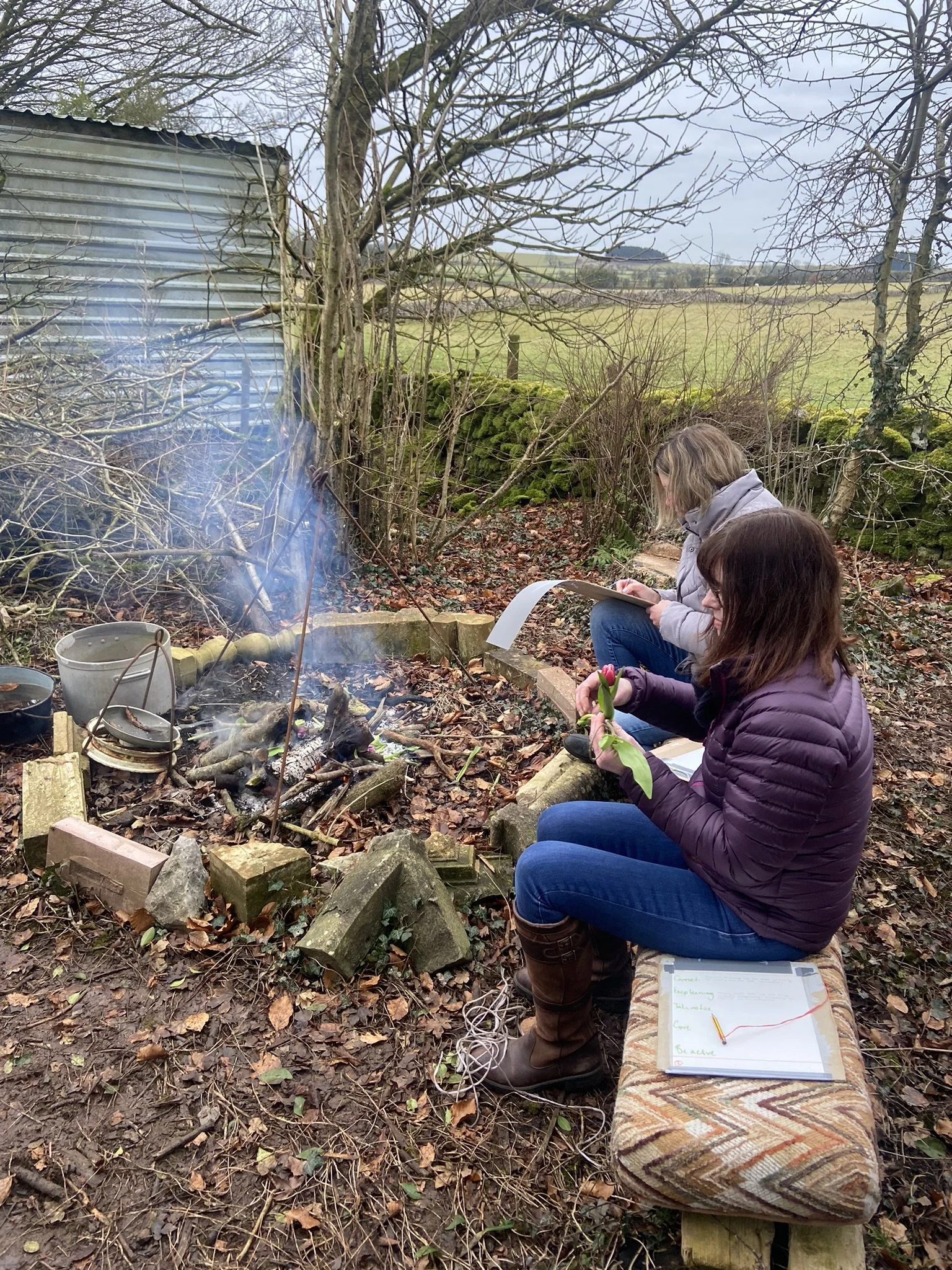 Two women sit outdoors near a small campfire with smoke rising, surrounded by leaf-covered ground, trees, and a stone wall in the background. One woman is holding tulips and a clipboard, while the other reads a book or notebook. They are dressed in w